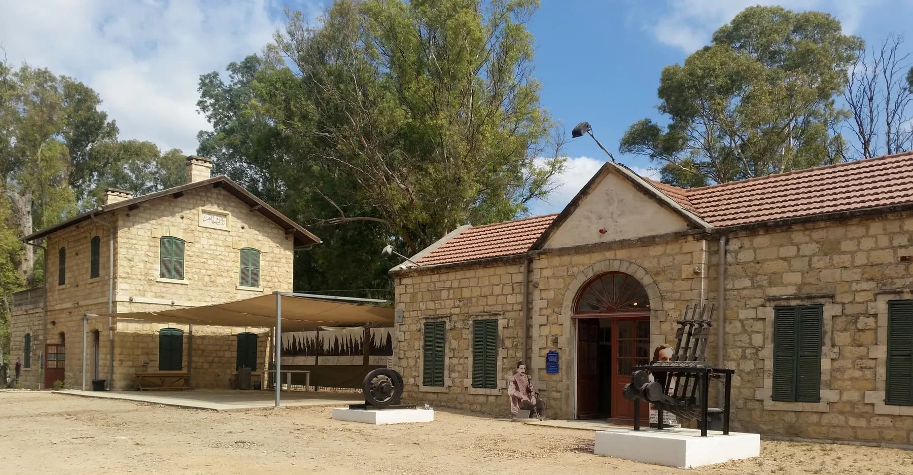 Old stone buildings with tiled roofs, green shutters and an arched doorway in Kfar Yehoshua, with old machinery displayed on white plinths in the sandy yard and tall eucalyptus trees behind.
