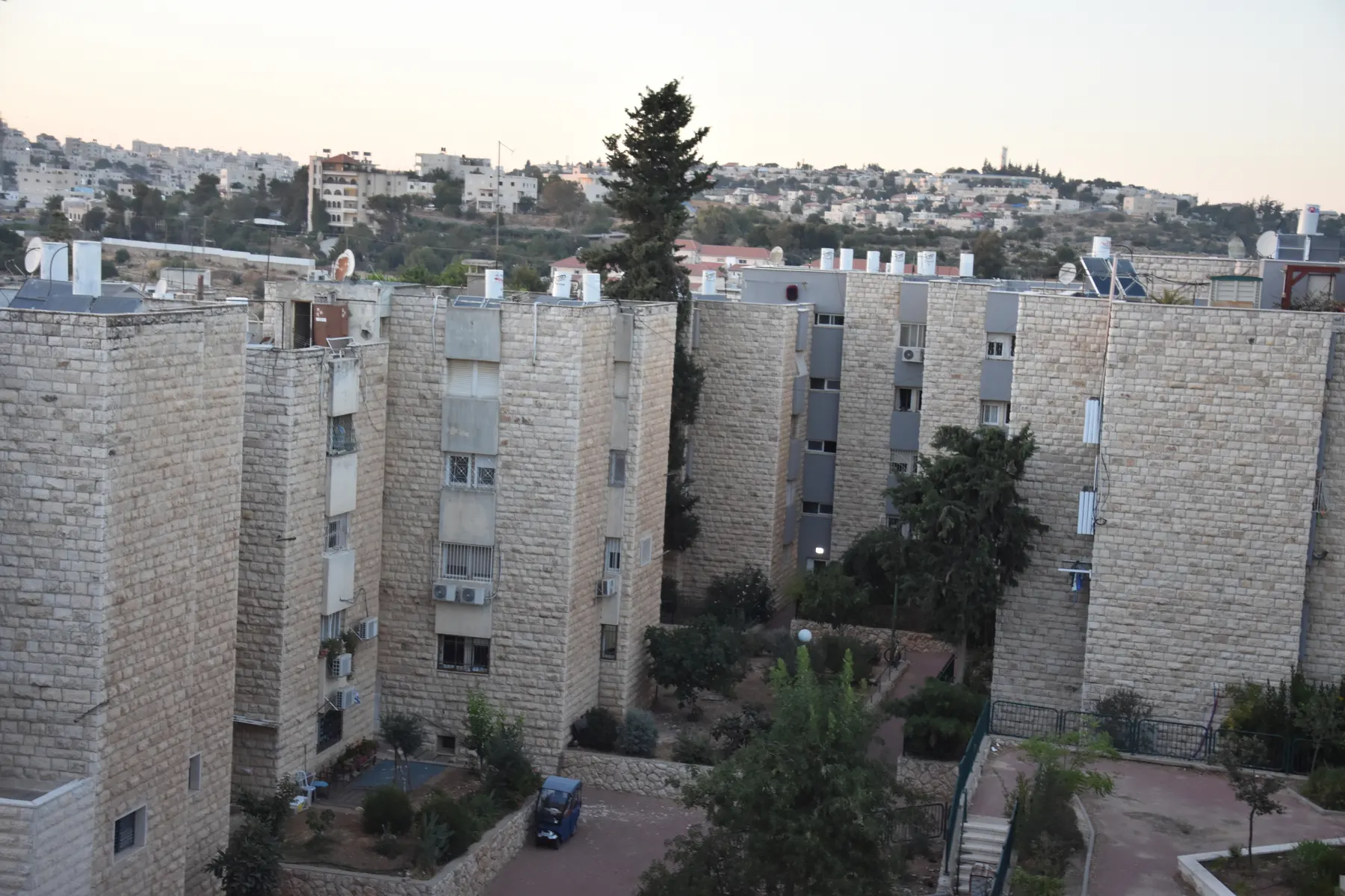 Stone-clad apartment blocks with gardens and trees between them in Kiryat Arba, with more houses on the hillside behind.