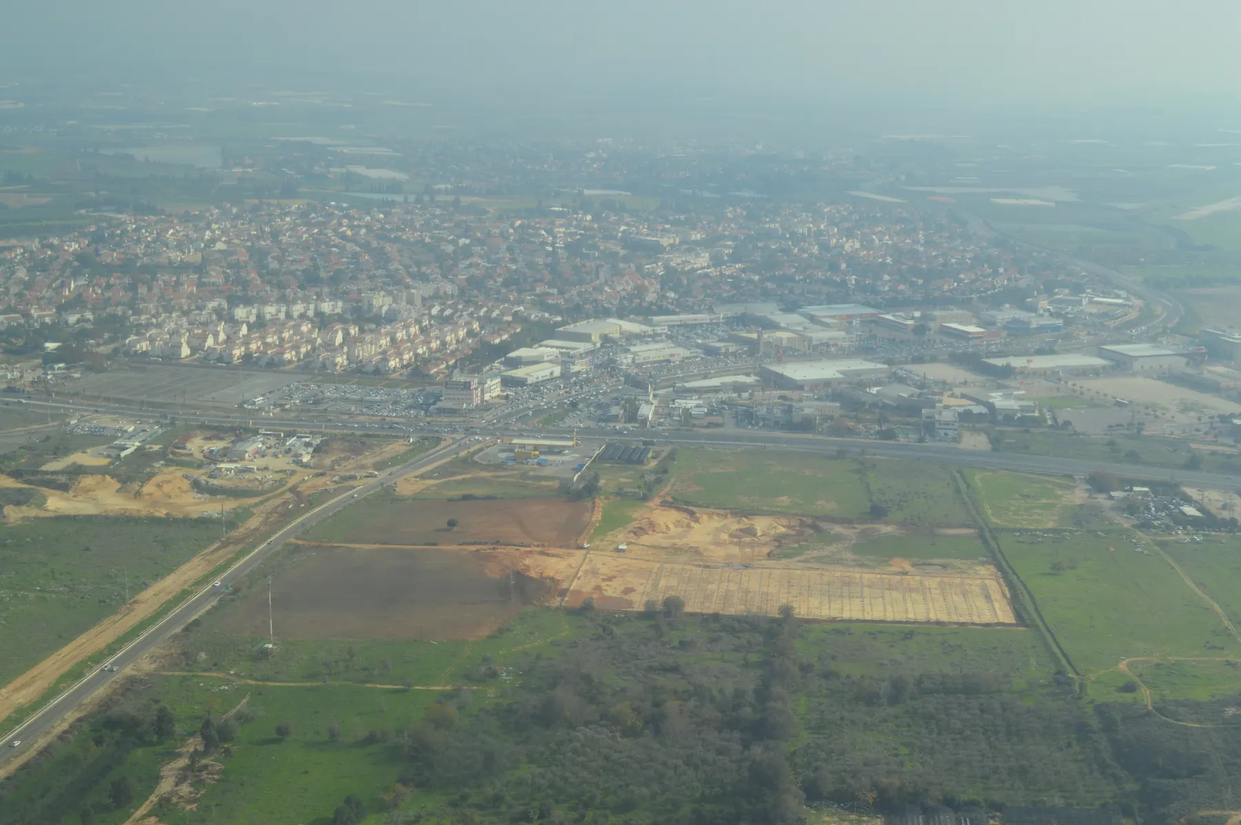 An aerial view of the houses of Kiryat Ekron beside a highway and a large commercial area, with fields in the foreground.
