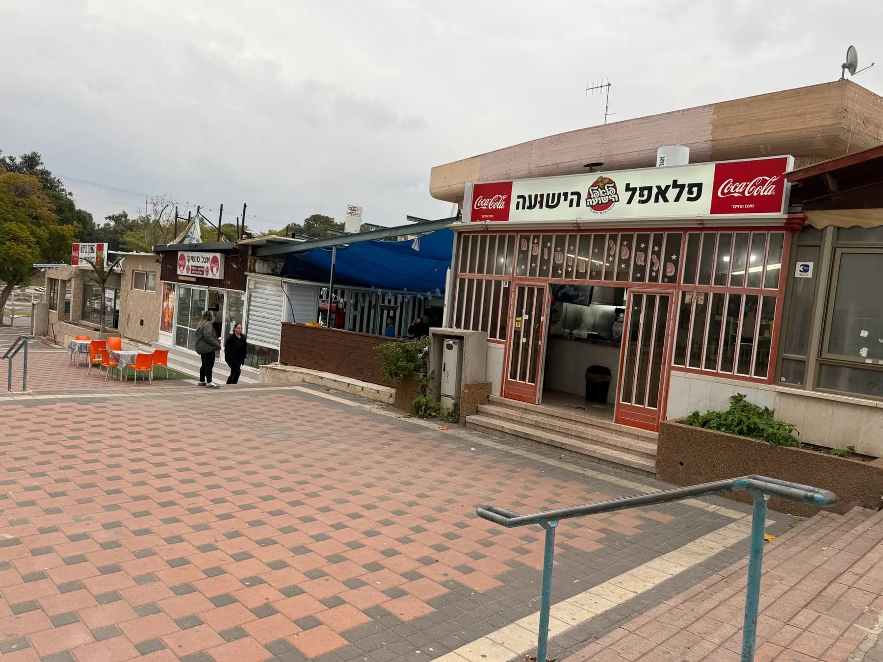 A paved square of small food stalls in Kiryat Gat, with a falafel shop and outdoor tables.