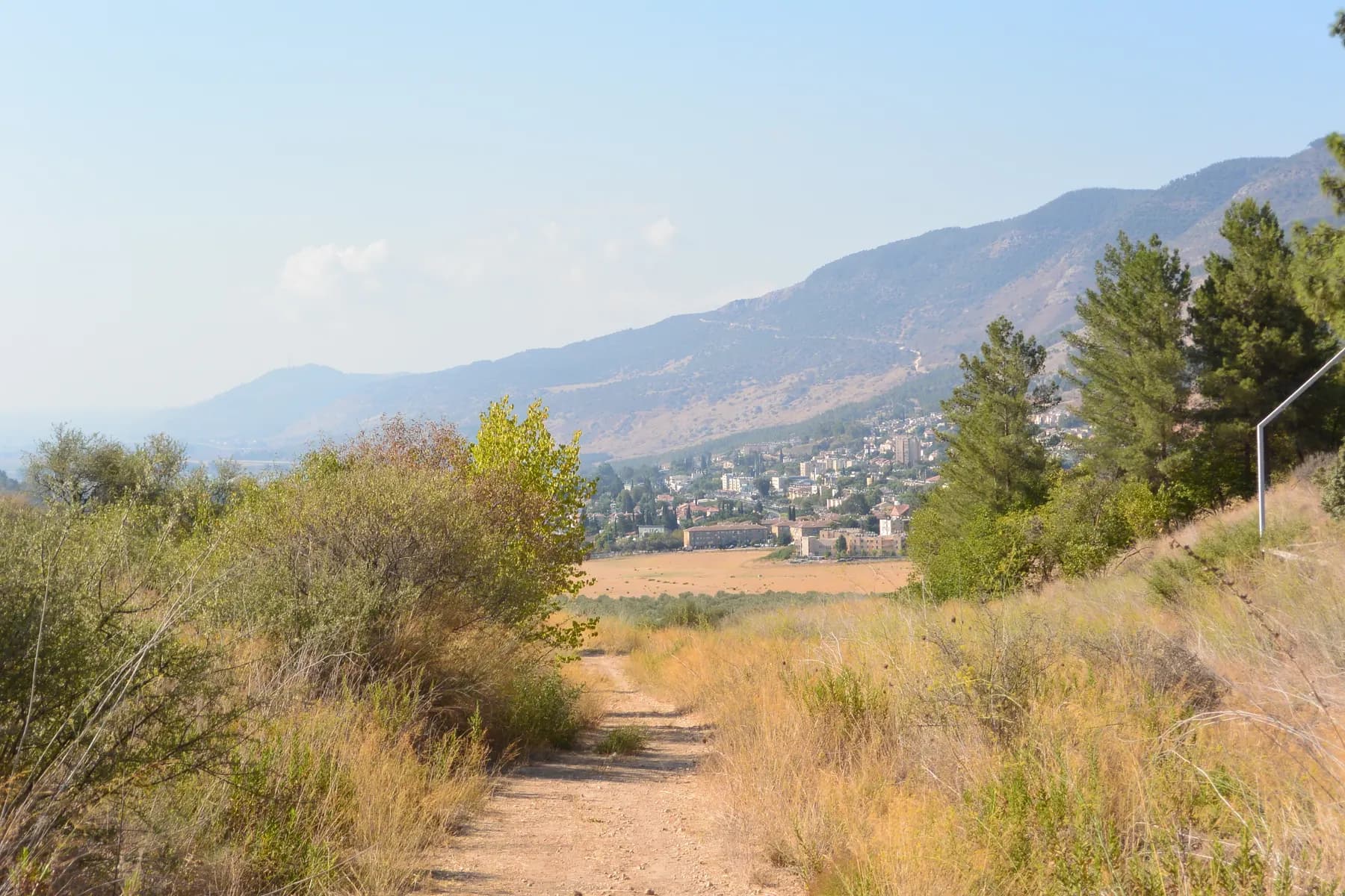 A dirt path through dry grass and bushes looking toward Kiryat Shmona in the valley, with a mountain ridge behind.