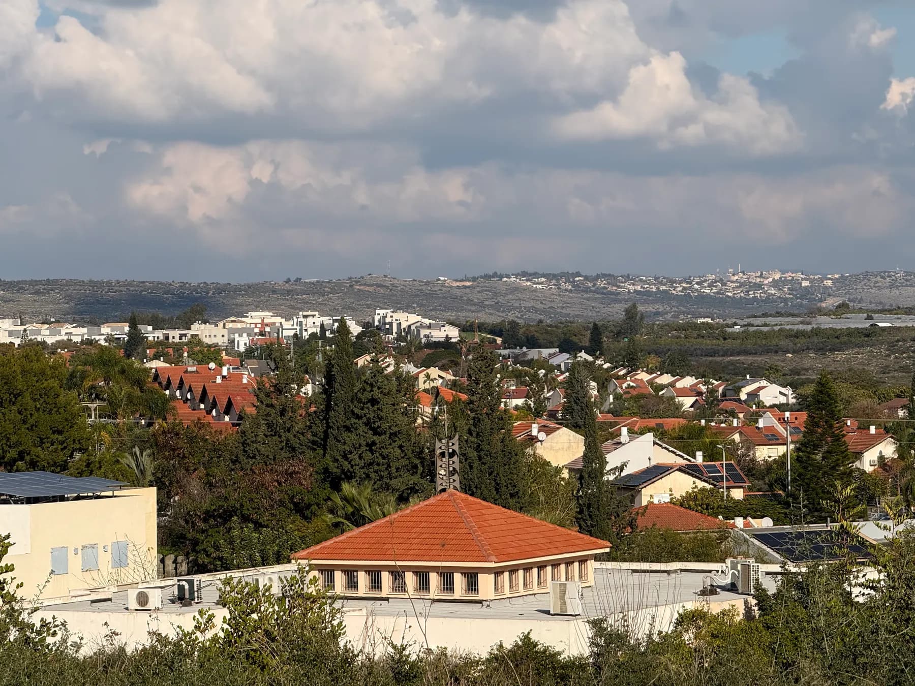 Red-tiled roofs and houses among trees in Kokhav Ya'ir-Tzur Yig'al, seen from a hilltop, with low hills and a village on the horizon.