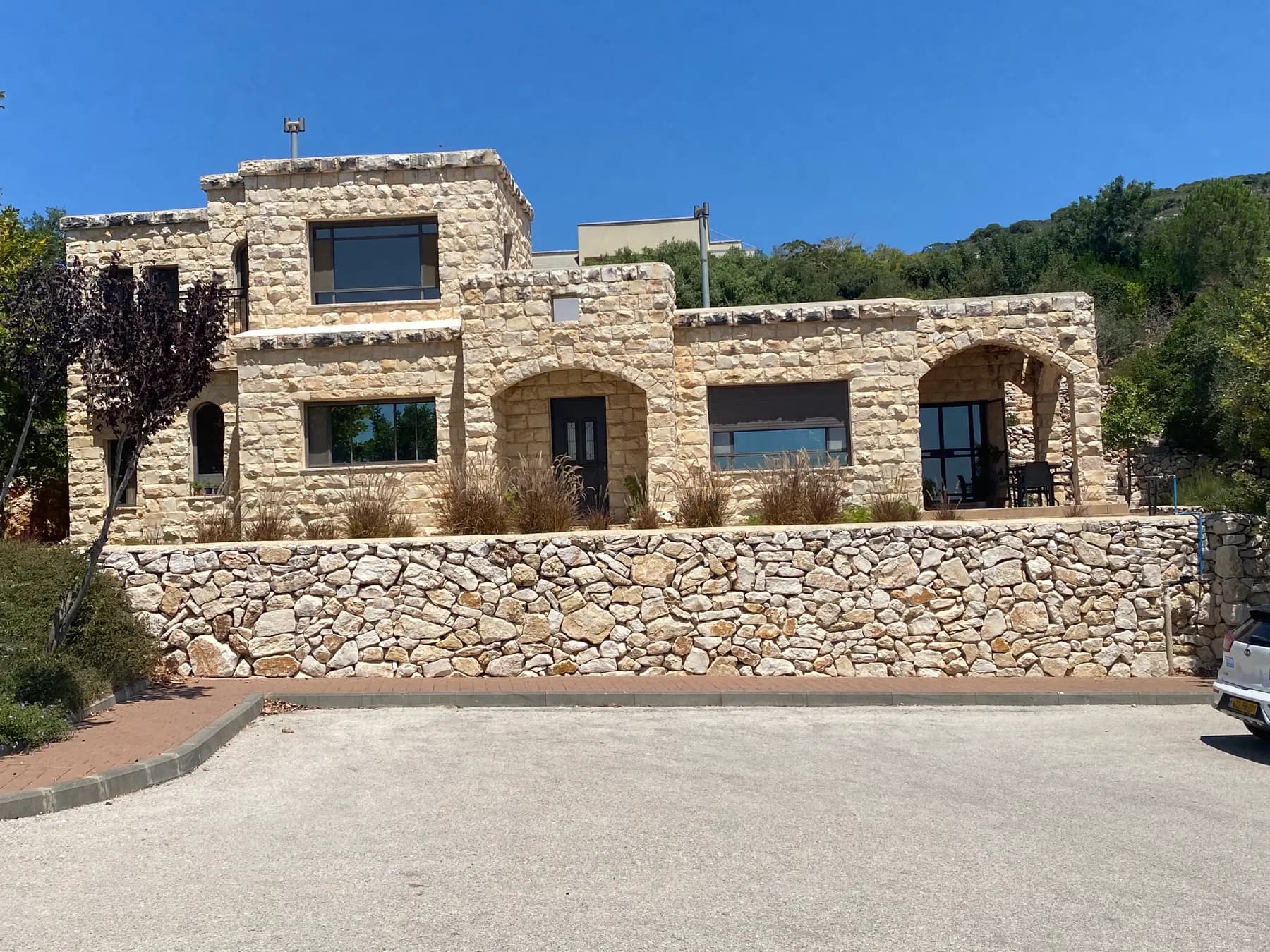 A two-storey stone house with arched openings above a stone retaining wall in Lavon, with green hills behind it.