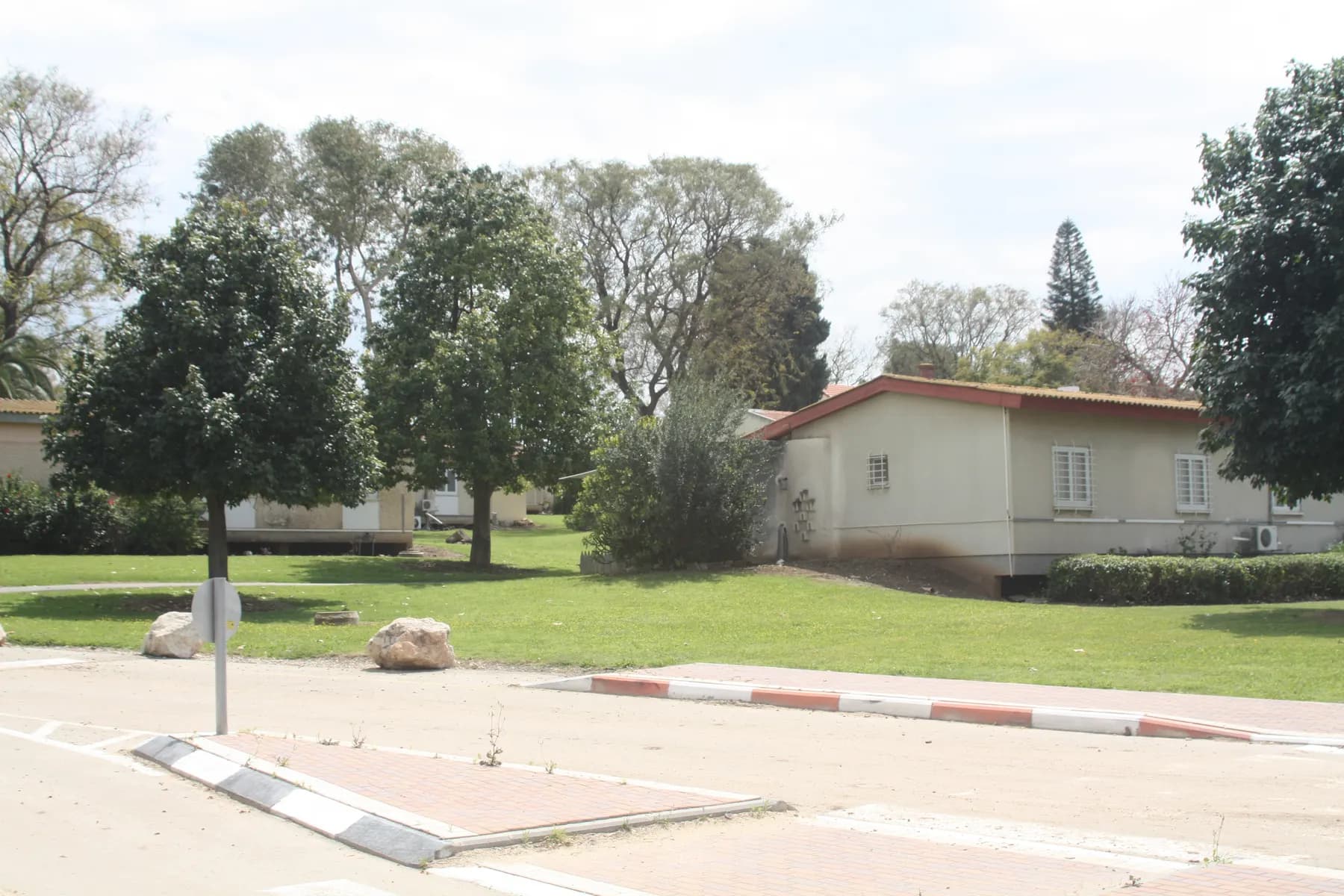 A single-storey house with a low tiled roof on a wide lawn among shade trees in Lehavot Haviva, beside a quiet road with a striped kerb.