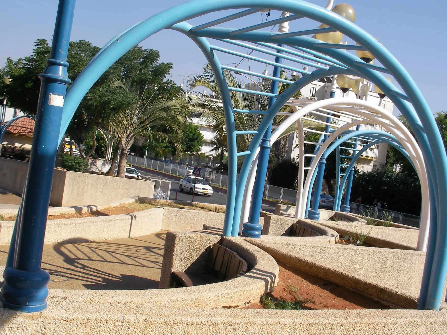Blue and white metal arches over a small plaza with built-in benches in Lod, with palms and apartment buildings behind.
