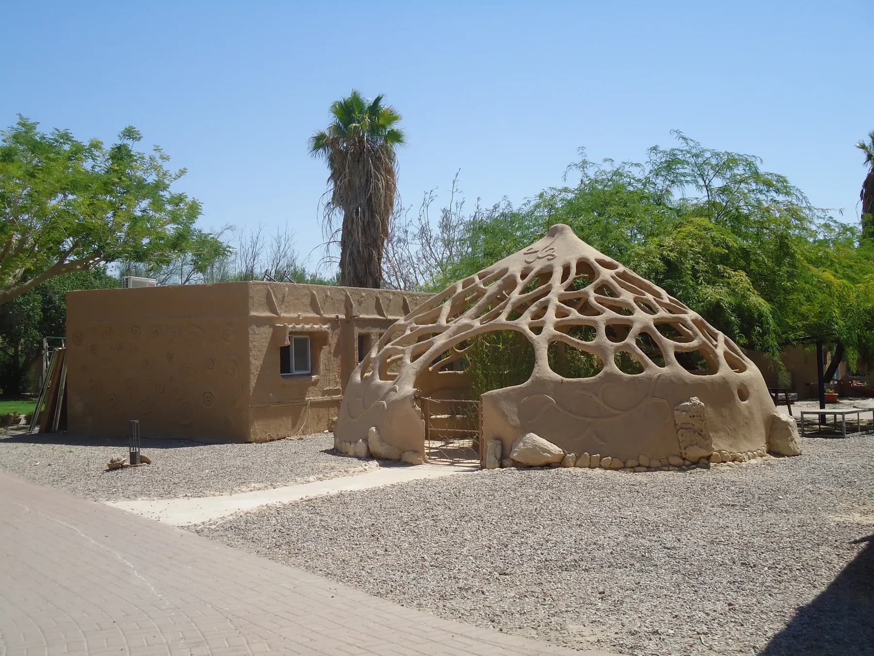 A mud-plastered house and a sculpted earthen dome with an open lattice roof among trees and palms in Lotan.
