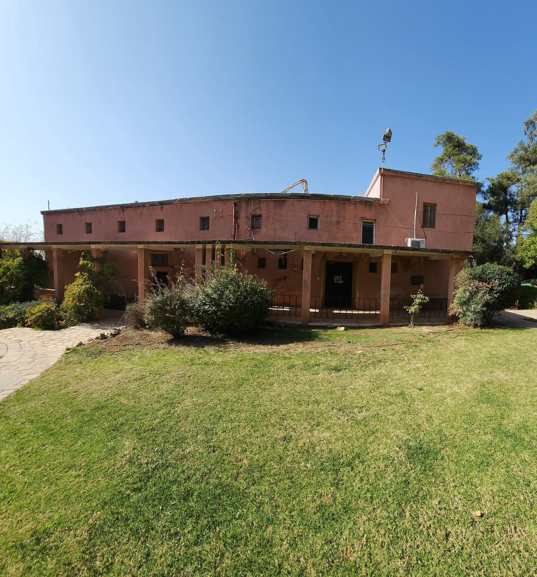 A long pink two-storey building with a covered veranda on pillars in Ma'ale HaHamisha, behind a lawn and shrubs.