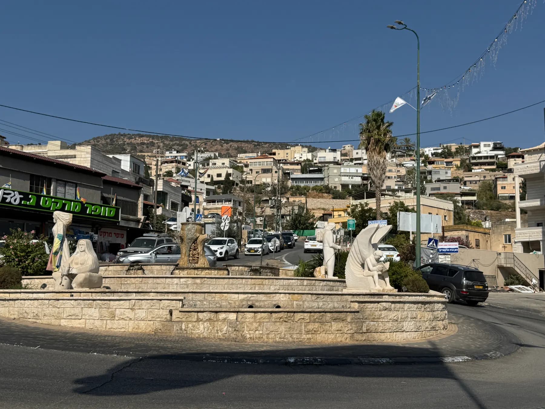 A roundabout with white stone statues and a fountain in Maghar, with houses climbing the hill behind.