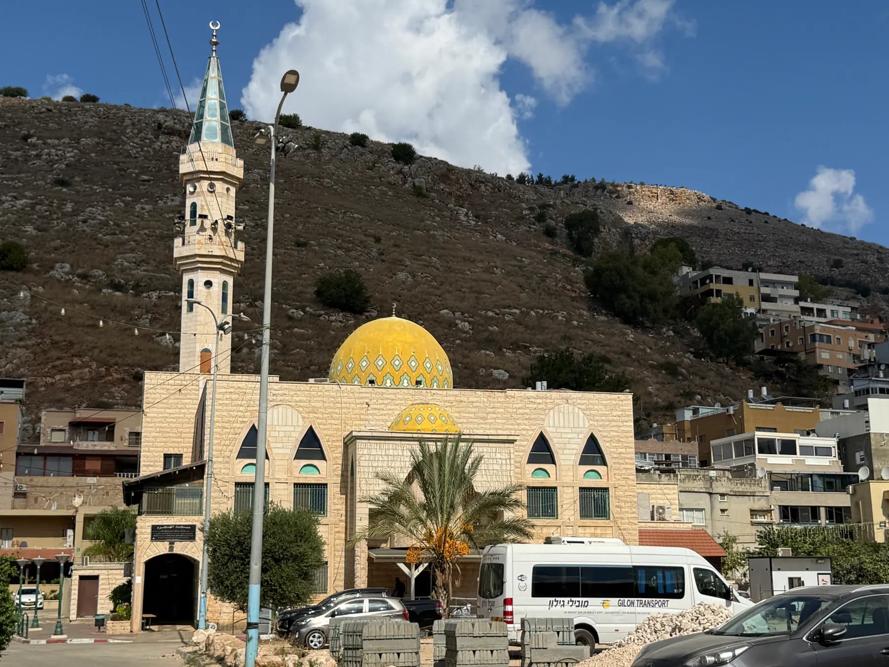 A mosque with a yellow dome and a tall minaret in Majd al-Krum, with houses climbing the rocky hillside behind.