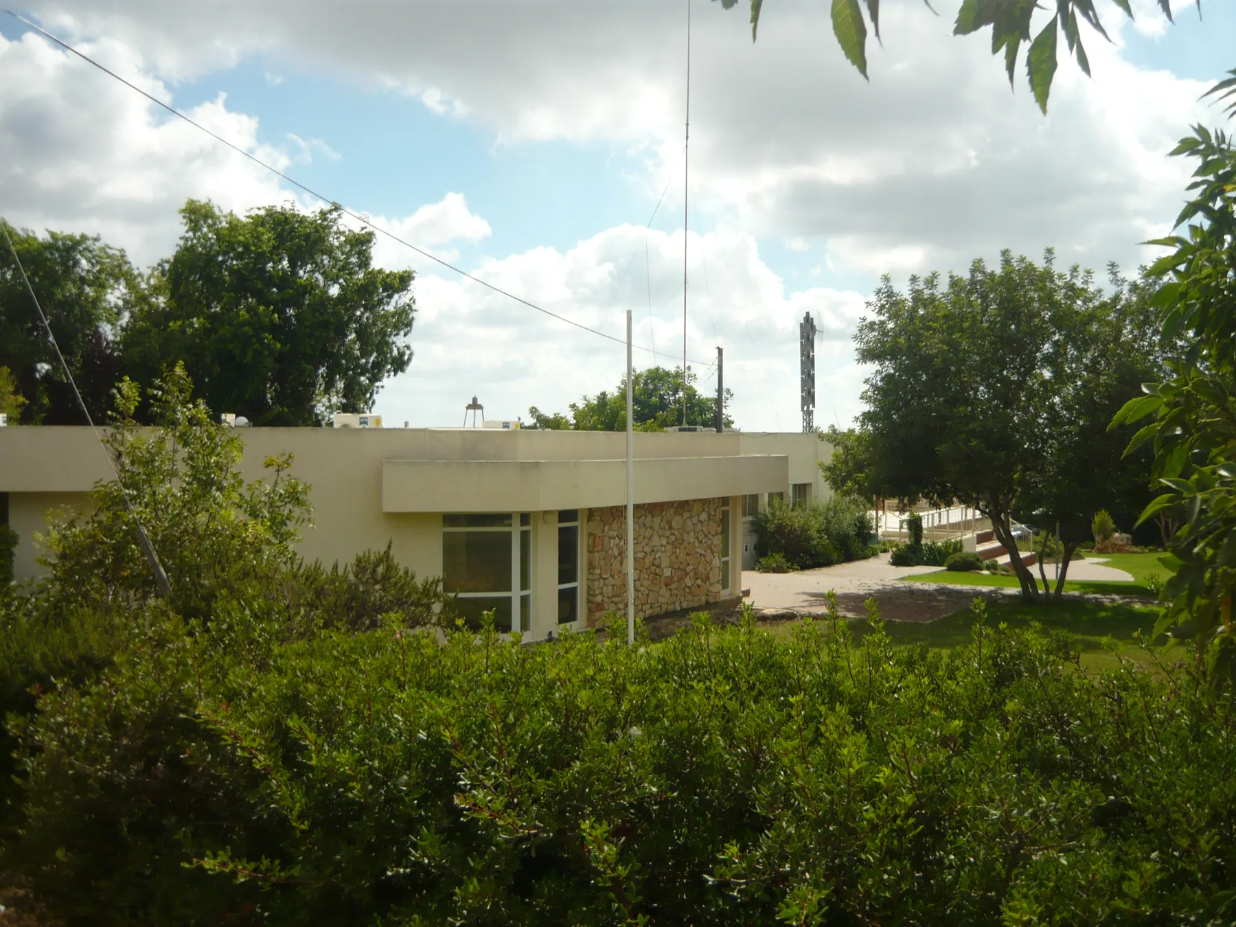 A low public building with a stone-clad wall behind green bushes and trees in Manof, under a cloudy sky.
