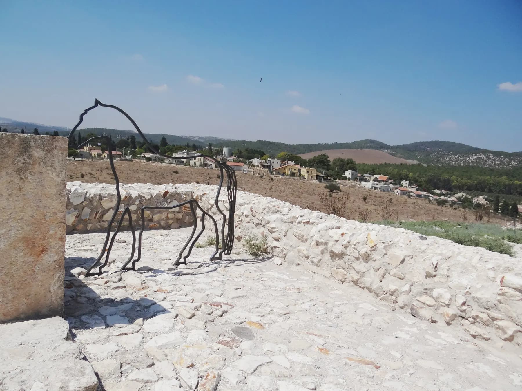 Houses and a water tower of Megiddo among trees across a dry field, seen from low stone walls with a wire horse sculpture in front.