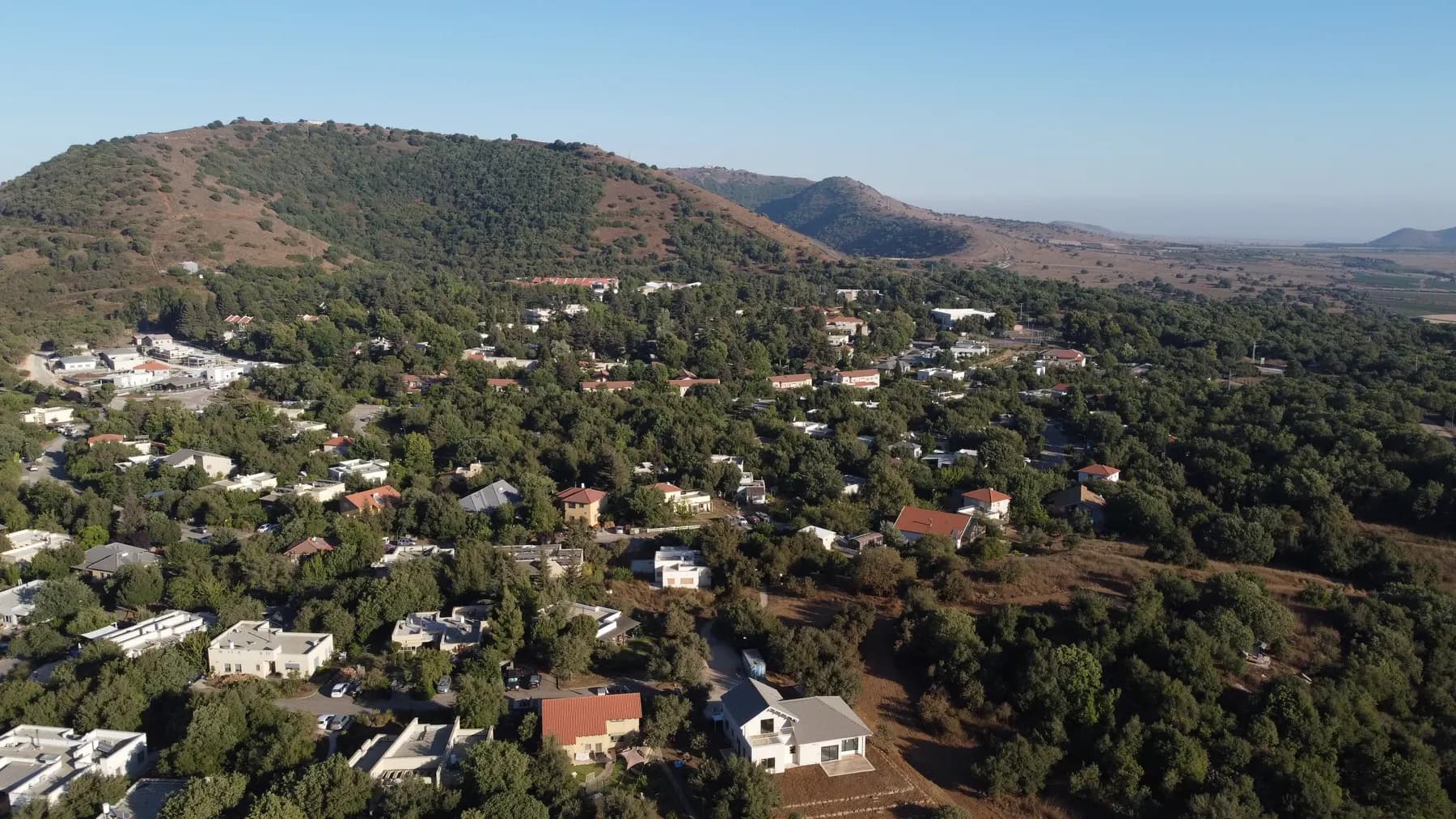 An aerial view of Merom Golan, its houses scattered among trees at the foot of a wooded hill, with open country stretching to the horizon.