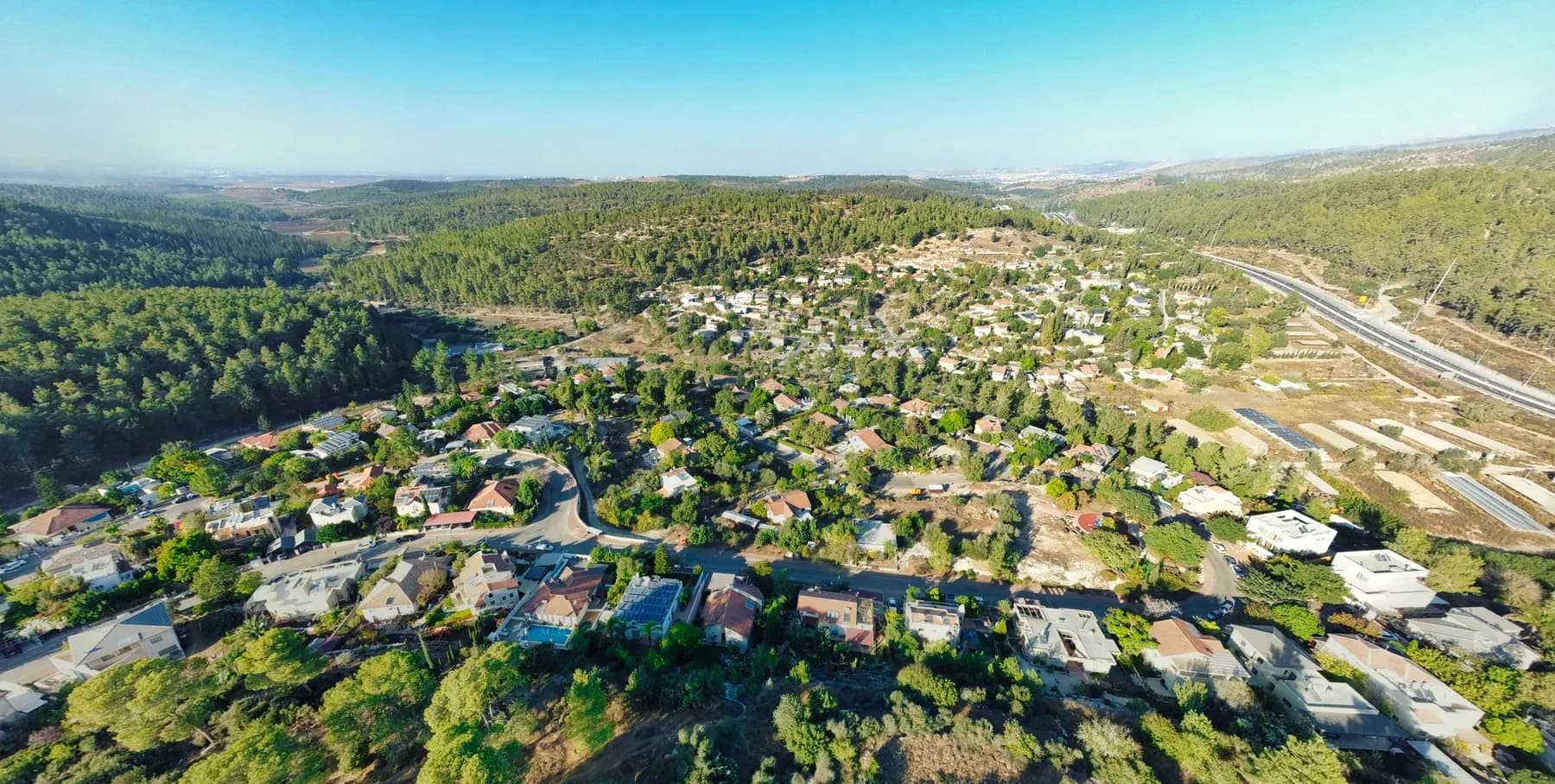 An aerial view of Mesilat Zion, its houses among trees across a hillside, with pine forest around it and a highway on the right.