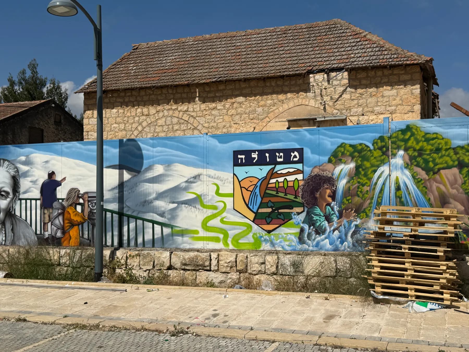 A painted mural of snowy mountains, a waterfall and the Metula emblem along a wall on the main street of Metula, with an old stone house with a tiled roof behind it.