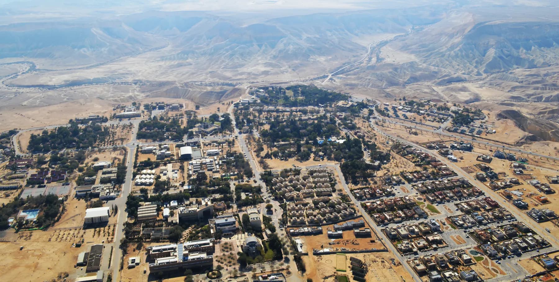 An aerial view over the streets, houses and trees of Midreshet Ben-Gurion, with a desert canyon and hills beyond.