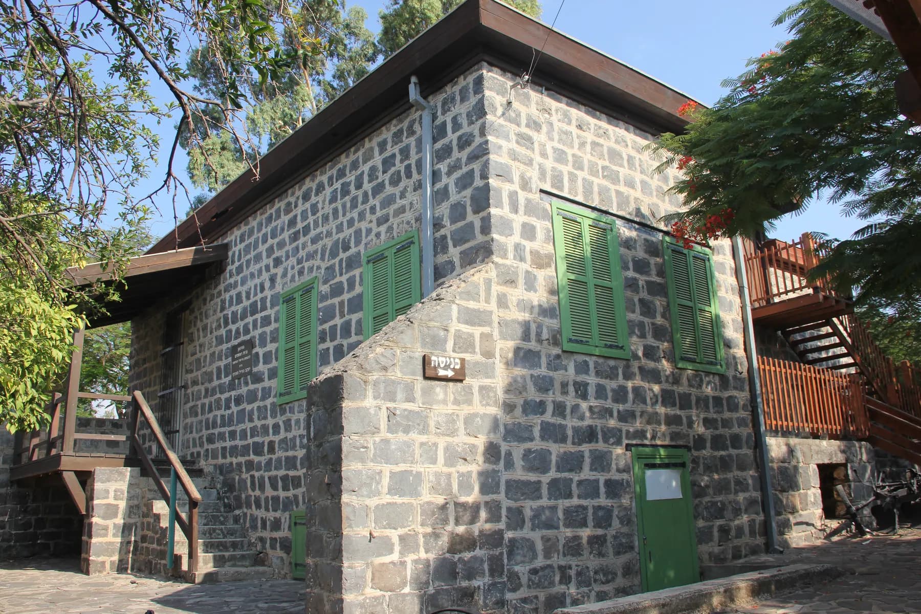 An old two-storey house of dark basalt stone with green shutters and a wooden outside staircase in Migdal, shaded by trees.