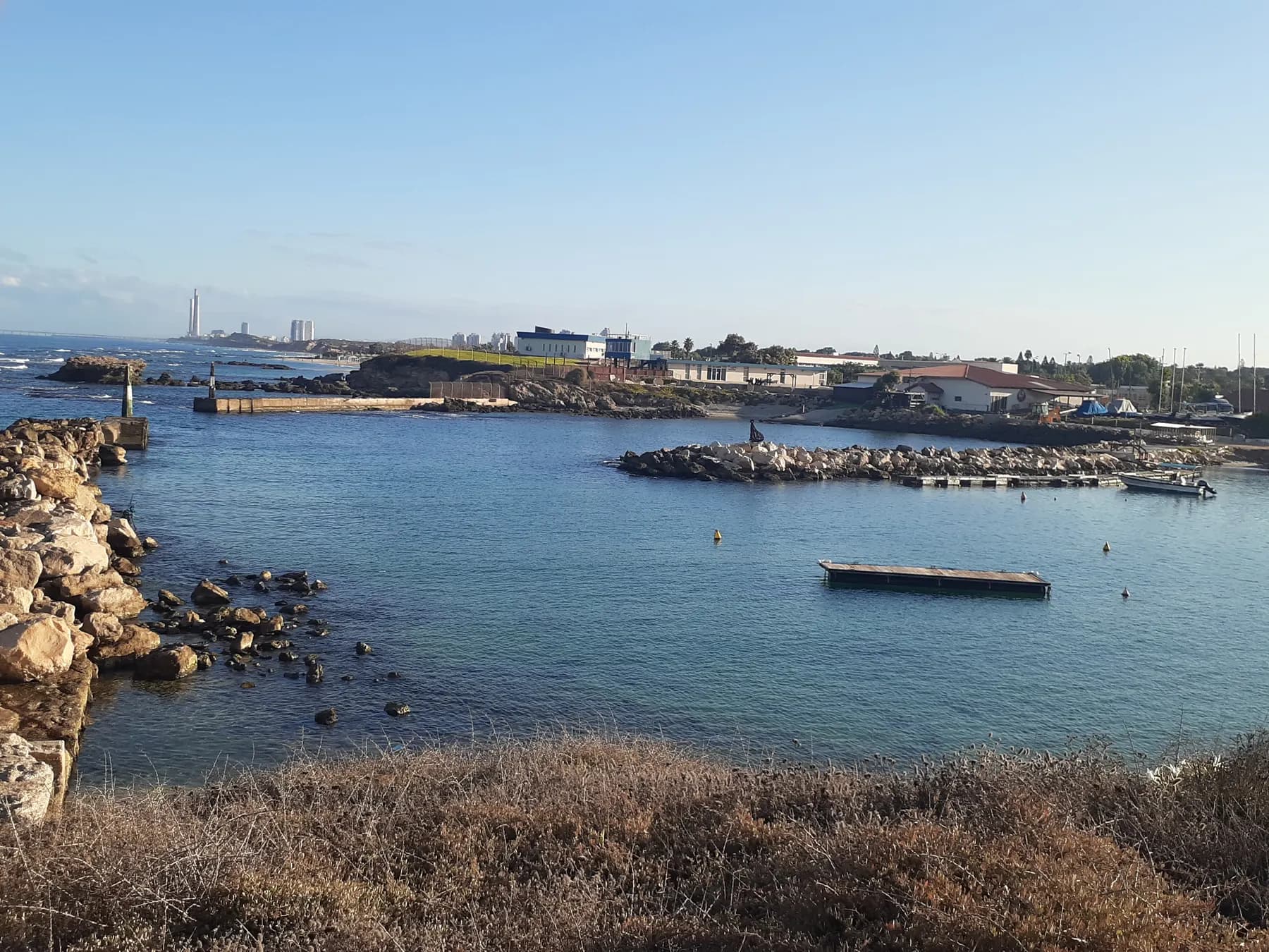 A small rocky bay at Mikhmoret with a floating jetty and a moored boat, and low buildings along the shore.