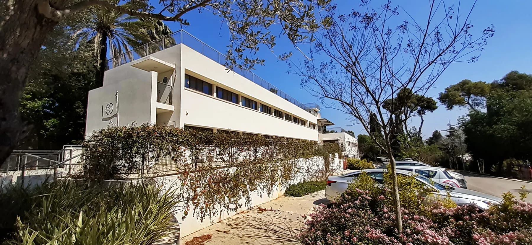 A long white modernist building behind flowering shrubs and trees in Mishmar HaEmek, under a blue sky.