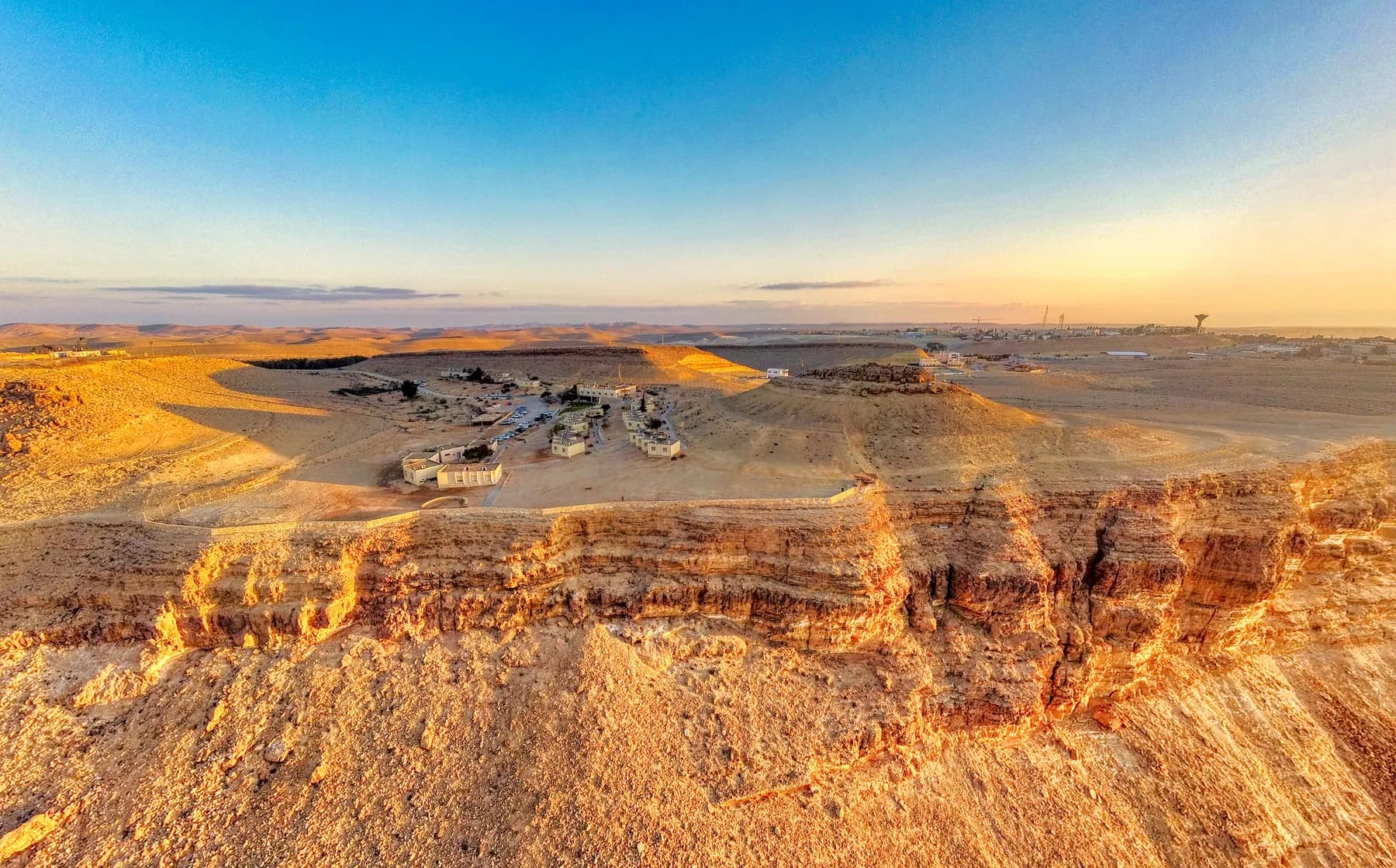 Low buildings on the rim of the Ramon Crater at Mitzpe Ramon, above a layered rock cliff lit by the low sun.
