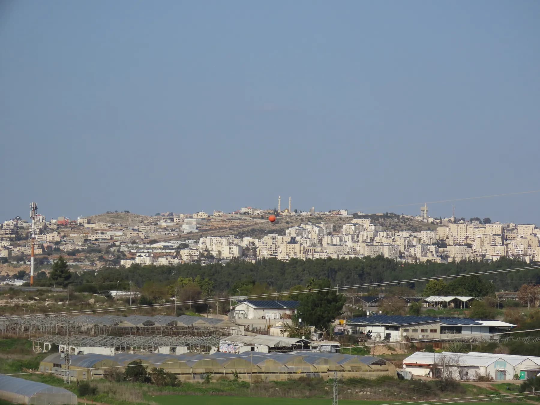 Apartment blocks of Modi'in Illit on a hillside, with greenhouses and farm buildings in the foreground.