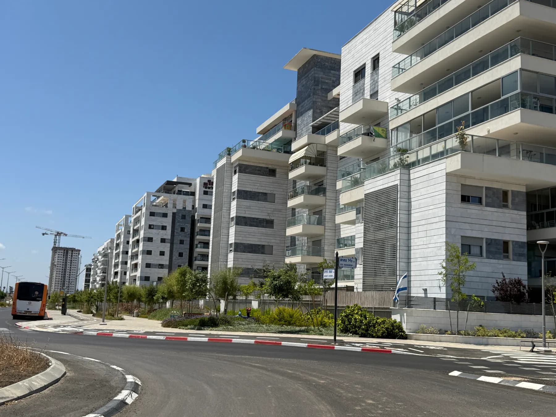 New apartment buildings along a street in the Yitzhak Shamir neighbourhood of Nahariya, with young trees and a bus.