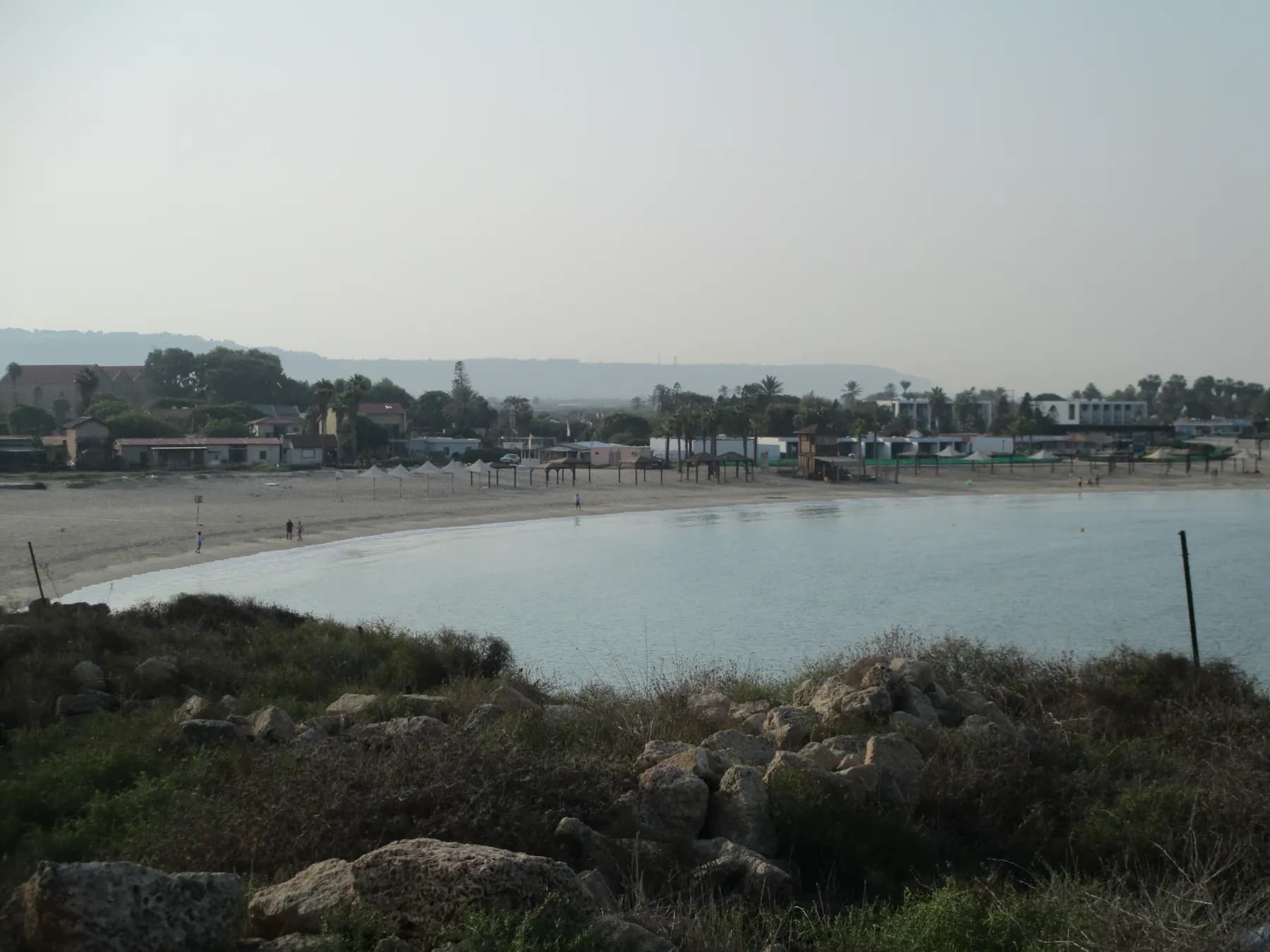 A curved sandy beach around a calm bay at Nahsholim, with the low buildings and palms of the kibbutz along the shore and a long ridge in the haze behind.