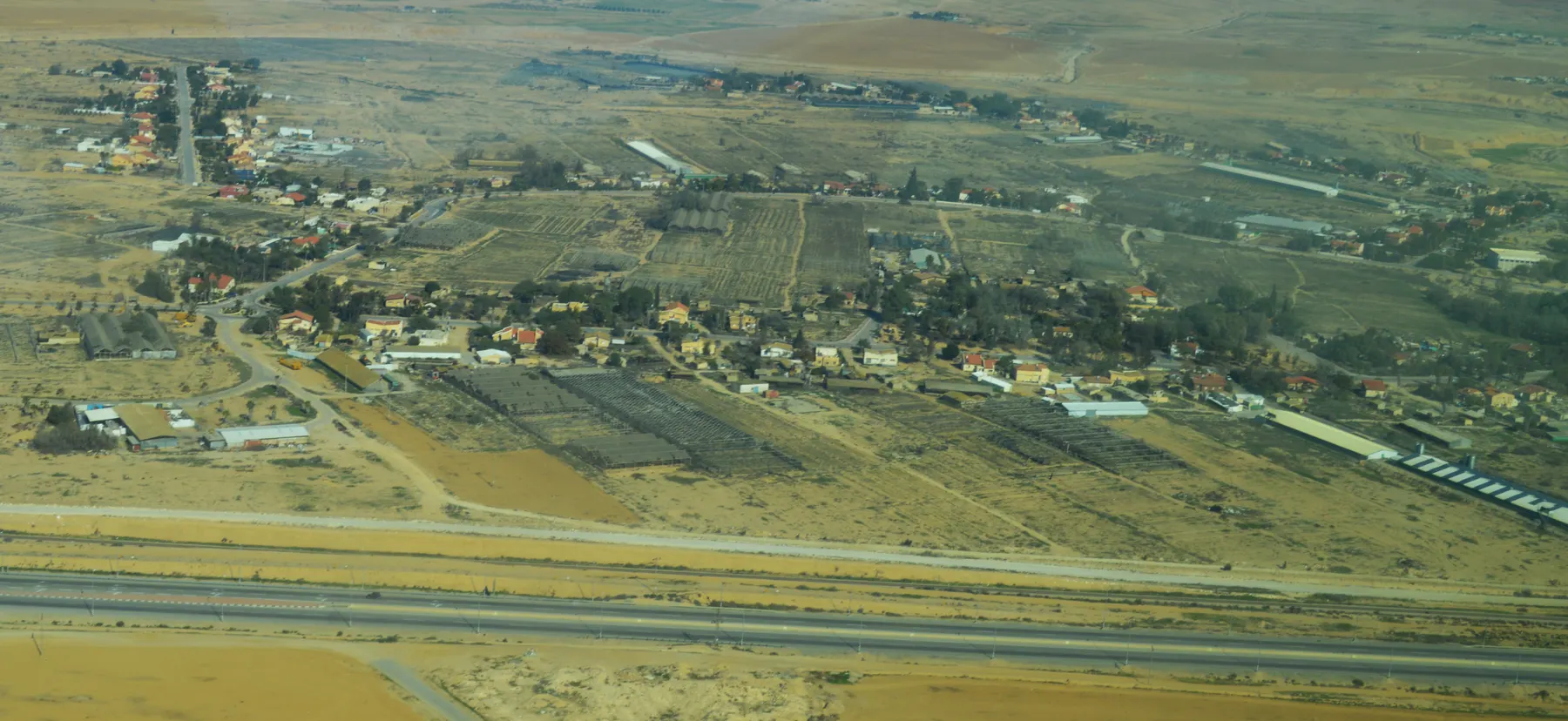 An aerial view of Nevatim, with houses and gardens along the edge of orchards and fields, and a divided road crossing the foreground.