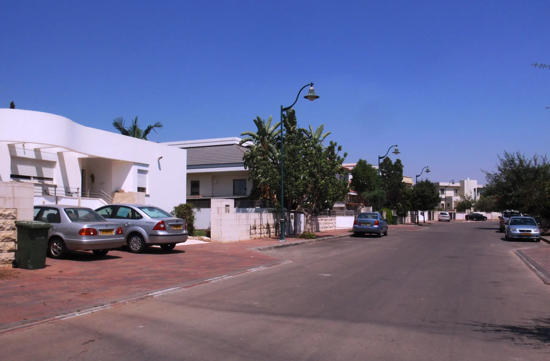 A quiet residential street in Neve Yarak with white houses, street lamps and young palm trees, and cars parked along the kerb.