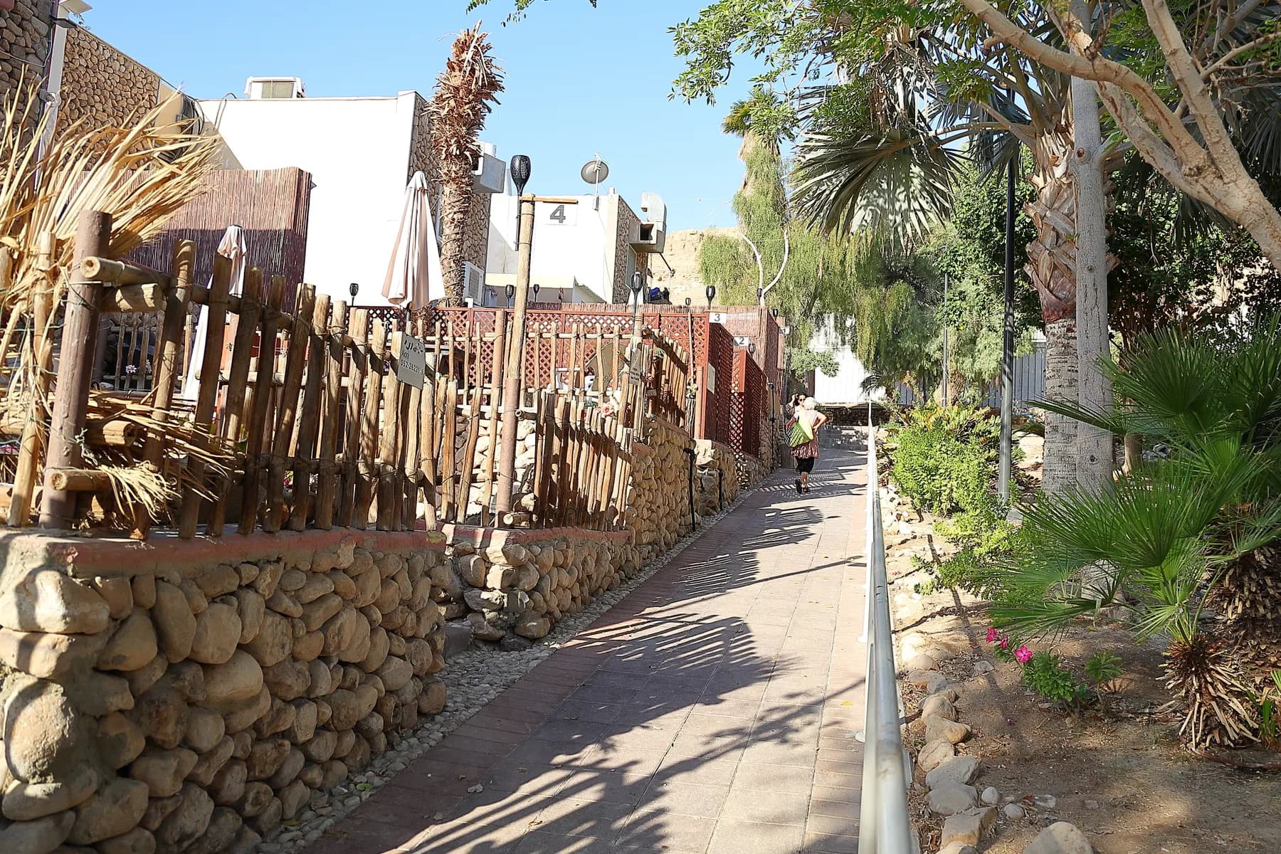 A paved lane lined with wooden fences, stone walls and palms in Neve Zohar, with white buildings and a railing alongside.