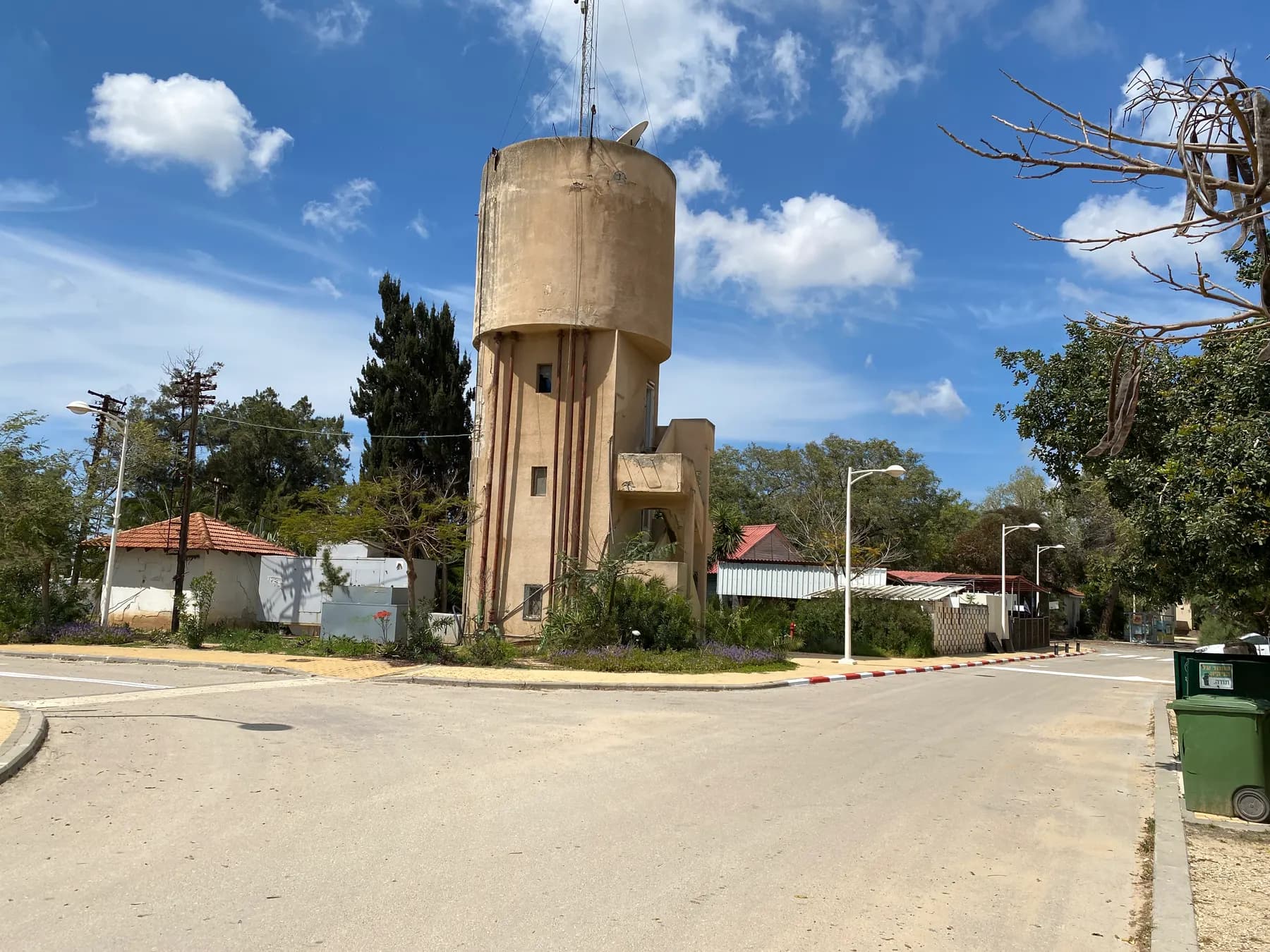 מגדל המים מבטון של ניר עם לצד כביש רחב, עם בתים בעלי גגות אדומים, עצים ופנסי רחוב תחת שמיים כחולים ועננים לבנים.