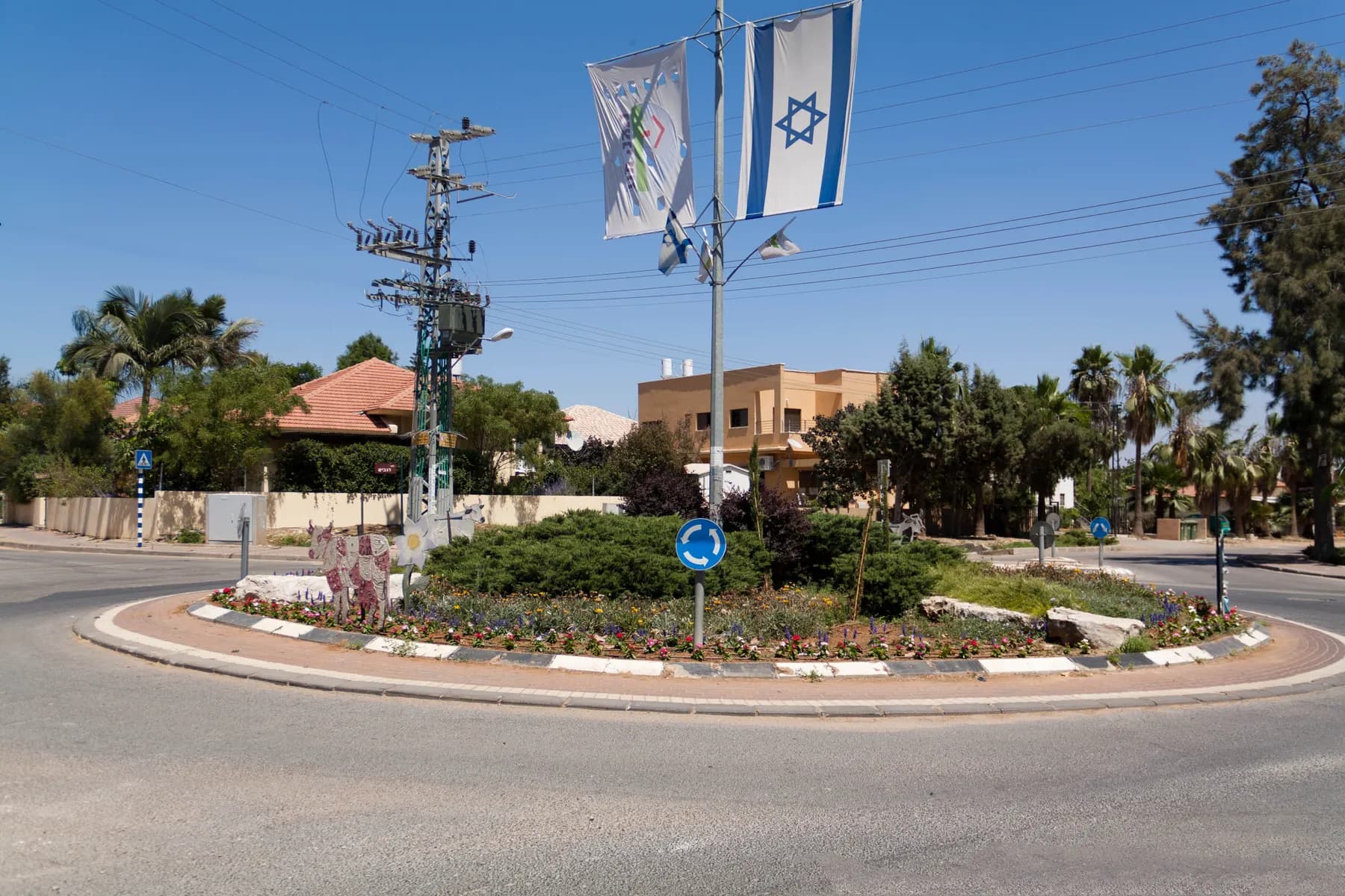 A roundabout planted with shrubs and flowers in Nitzanei Oz, with flags on a lamp post and red-roofed houses and palm trees behind.