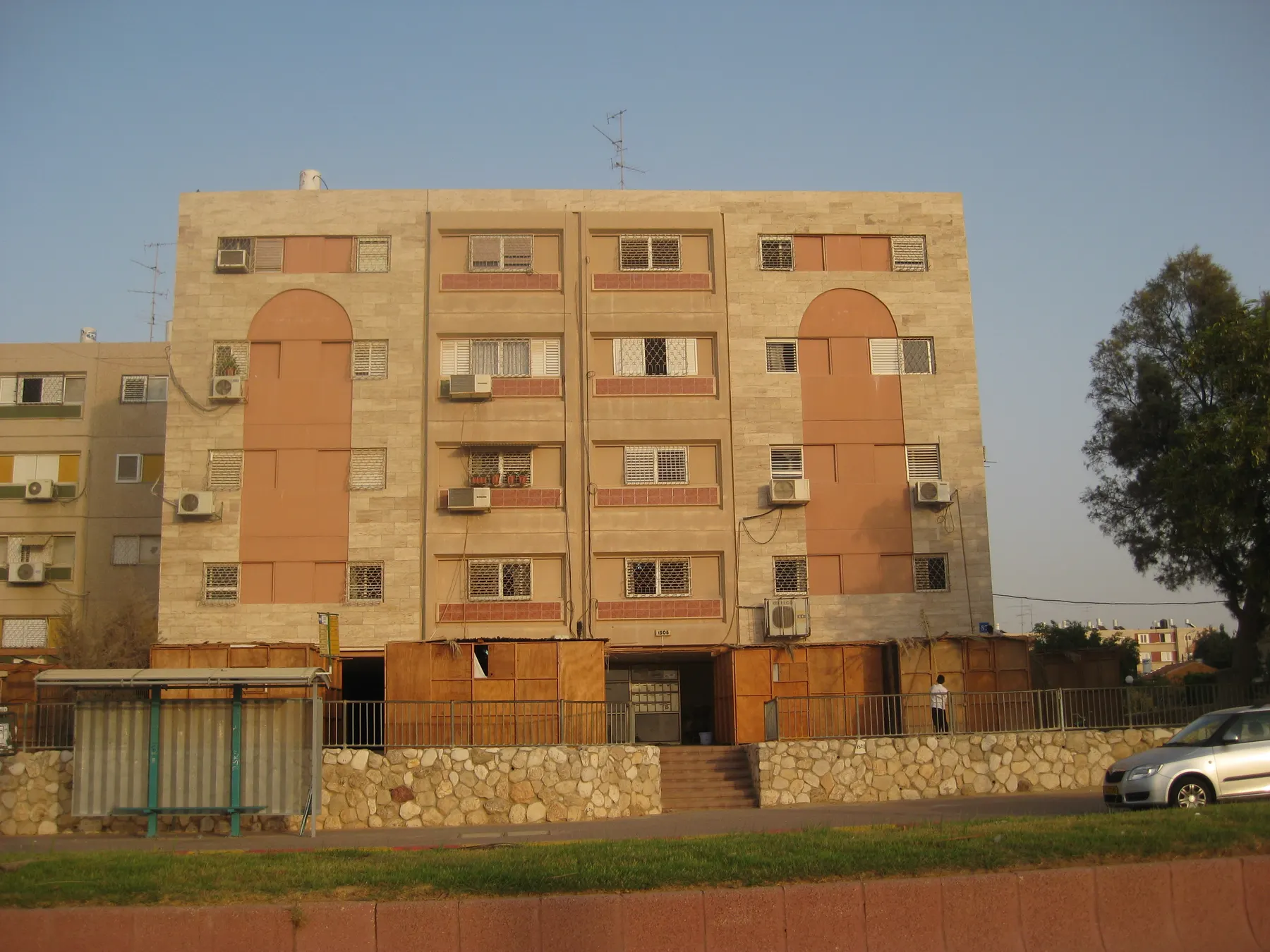 A four-storey apartment building with arched panels on its facade in Ofakim, with wooden sukkah booths in front, in warm evening light.