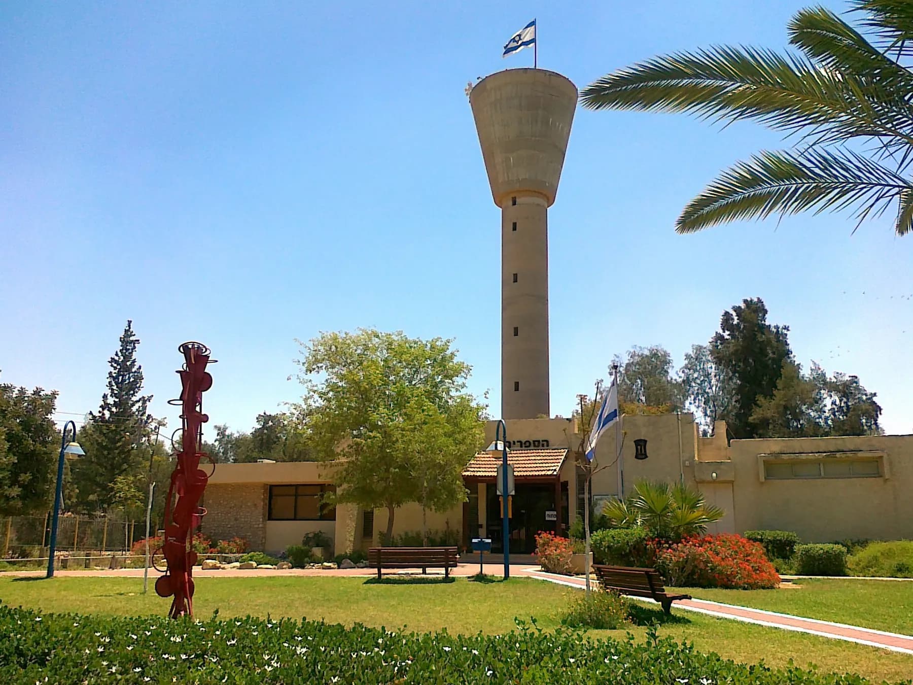 A tall water tower behind a low building with a flag in Omer, with a lawn, benches and a red sculpture in front.