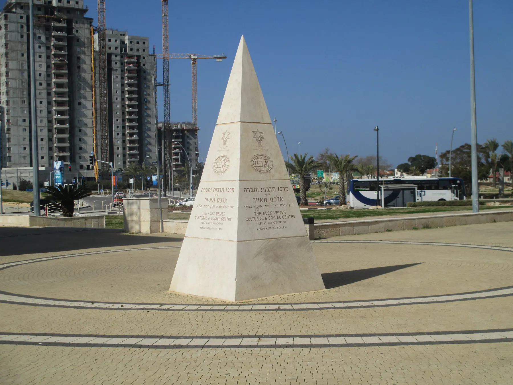 A white stone dedication pyramid on a paved square in Or Akiva, with new apartment towers and cranes behind.