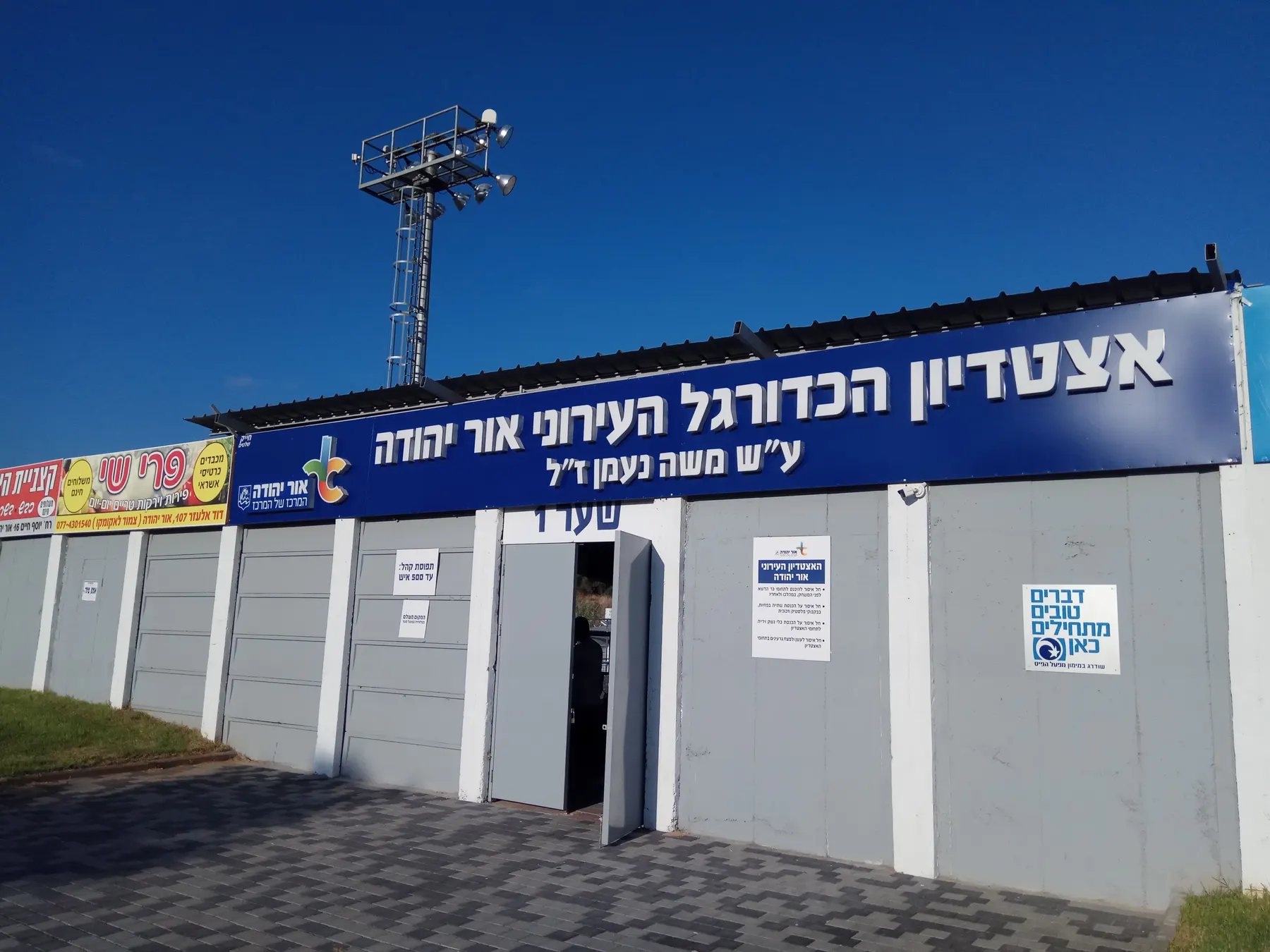 The gates of the Naaman football stadium in Or Yehuda, under a long blue sign, with a floodlight mast behind.
