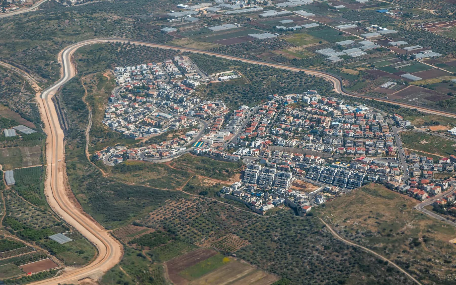 An aerial view of the red-roofed houses of the Beit Vagan neighbourhood in Oranit, ringed by a winding road, olive groves and fields.