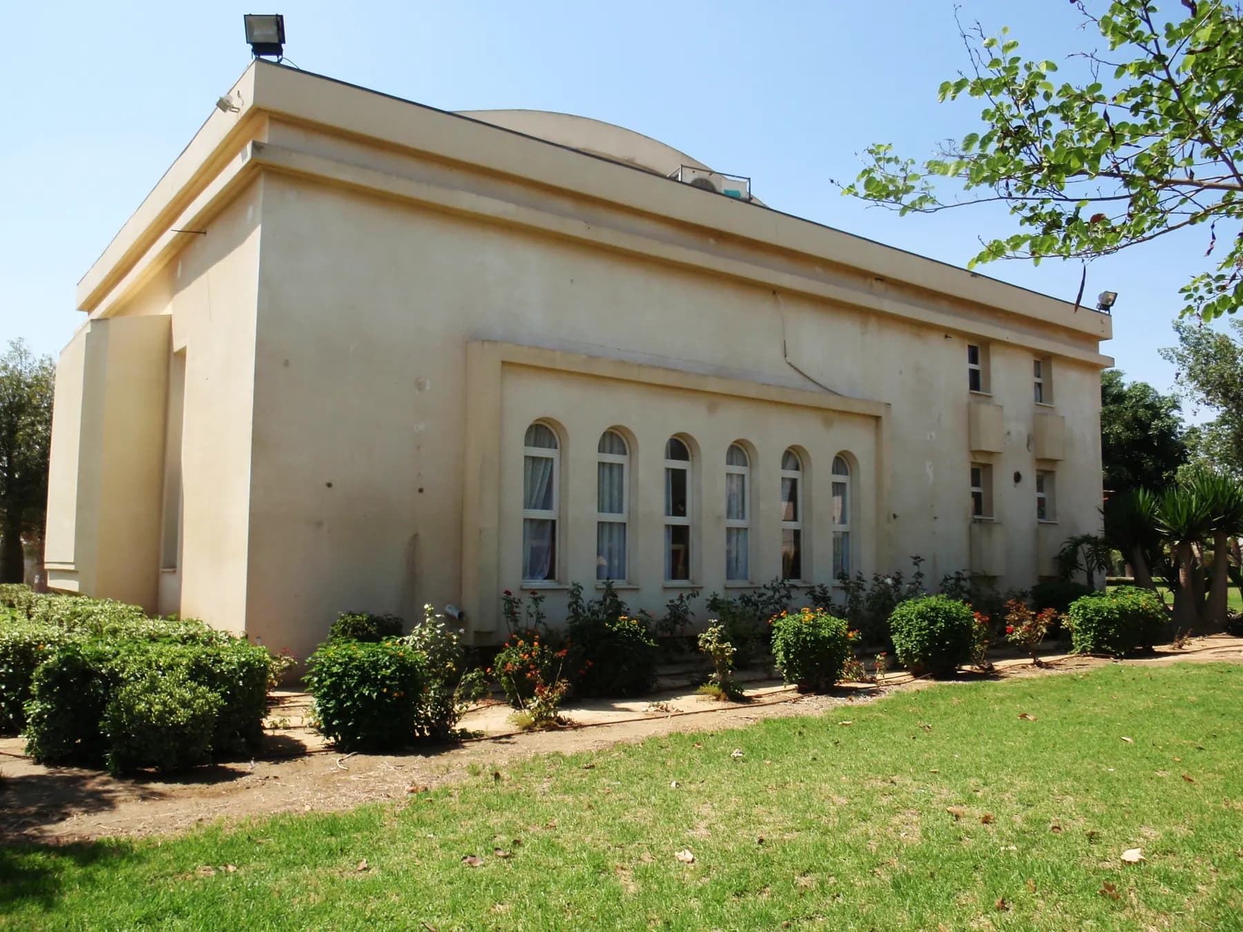 The synagogue of Pa'amei Tashaz, a large cream-coloured building with a row of tall arched windows, behind a lawn and rounded shrubs.