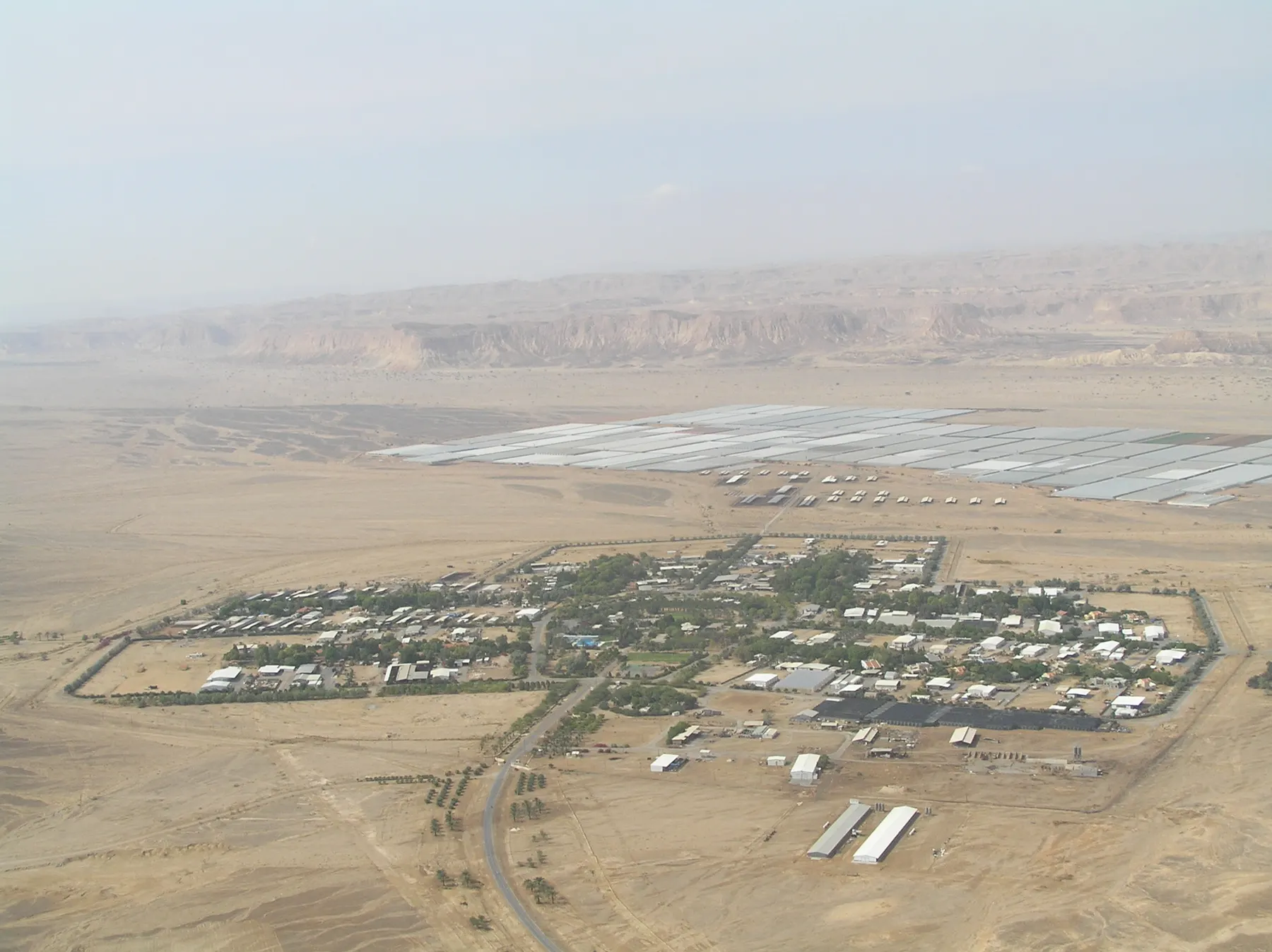 An aerial view of Paran, a fenced desert village among trees, with greenhouses and cliffs behind it.