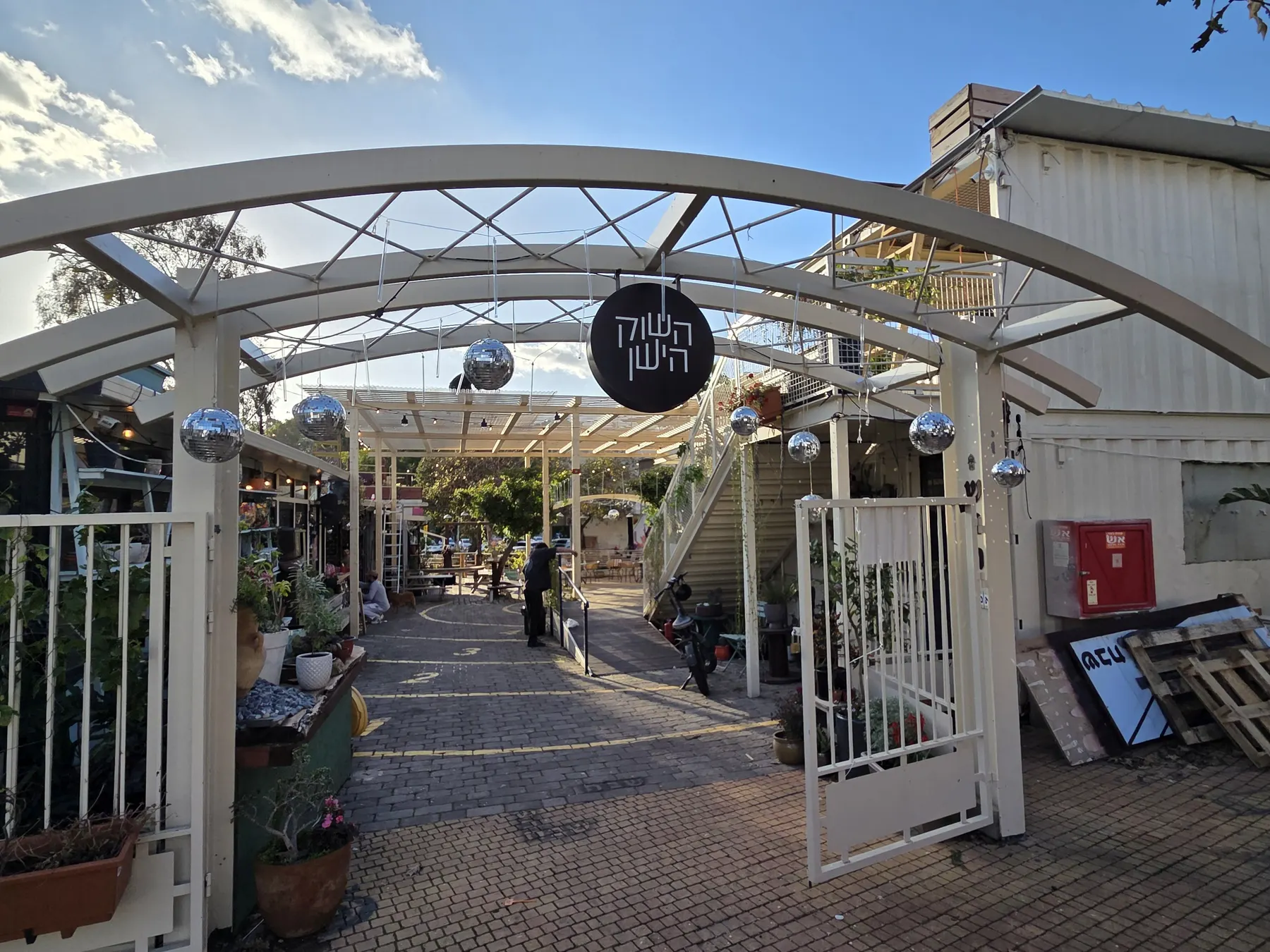 The gate into the old market of Pardes Hanna, an arch hung with mirror balls and a round sign reading HaShuk HaYashan, with plants along the alley