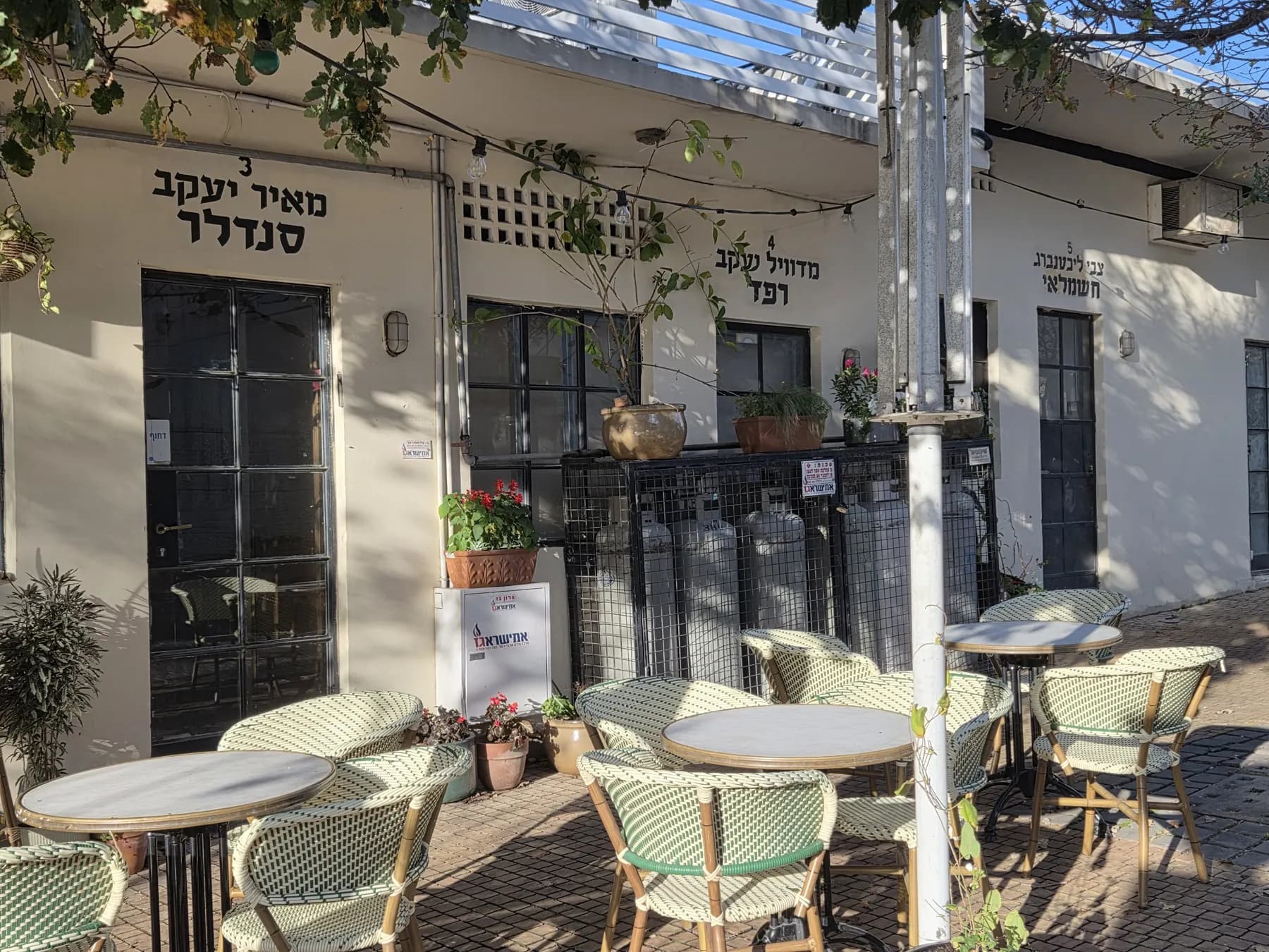 Bistro tables and rattan chairs under a pergola in front of cream-coloured shopfronts that still carry the old painted Hebrew signs of the Pardes Hanna market