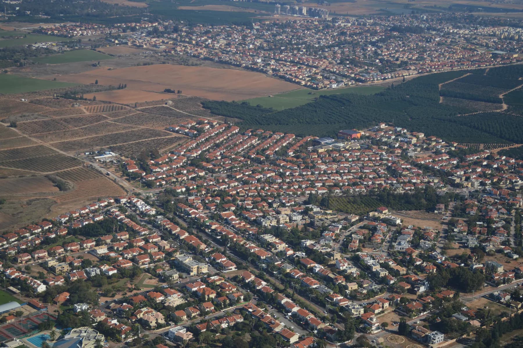 An aerial view over the red-roofed houses and tree-lined streets of Pardesiya, with orchards and fields beyond and a larger town on the horizon.