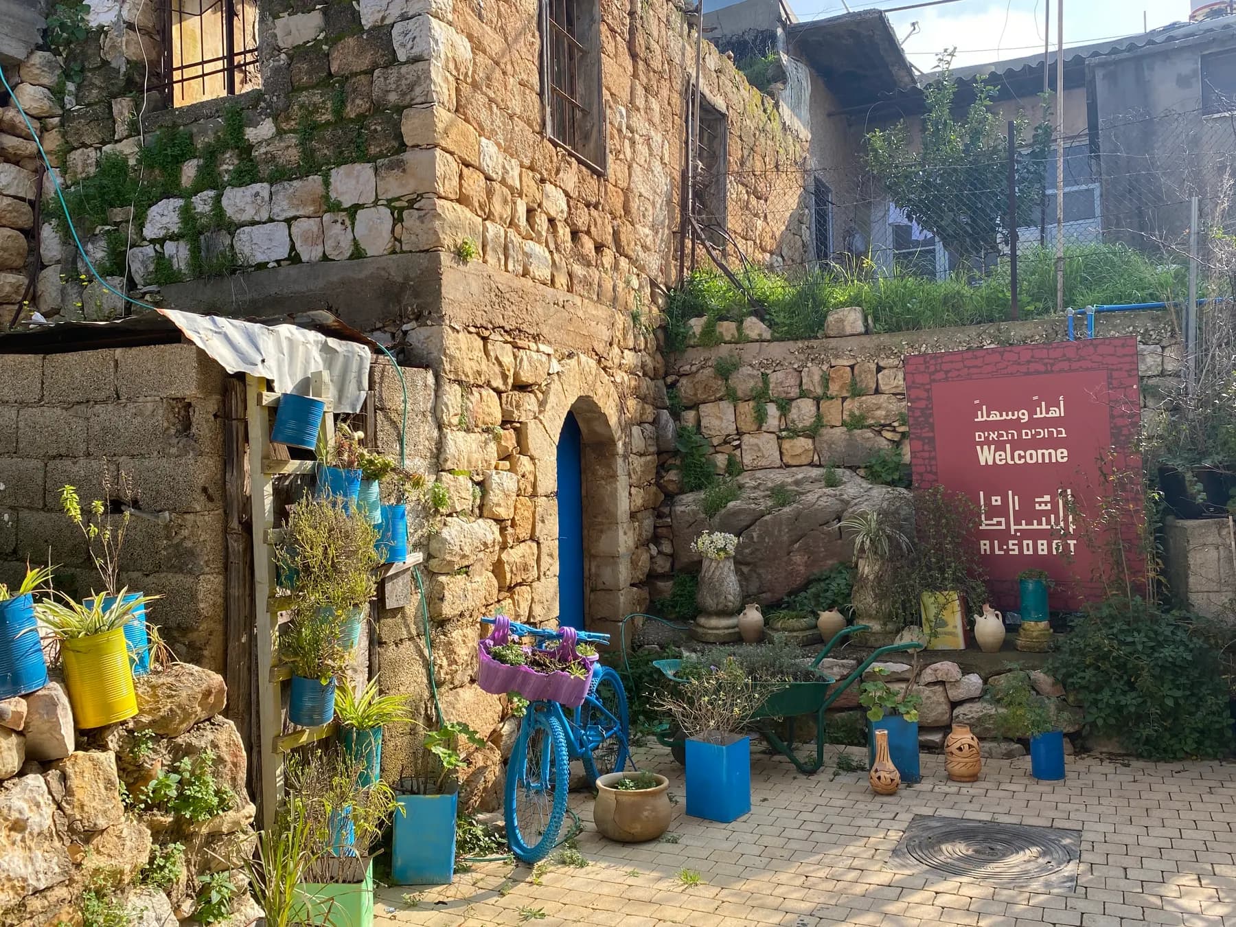 An old stone house with a blue door, plants in painted pots, a bicycle planter and a welcome sign in the old core of Rameh.