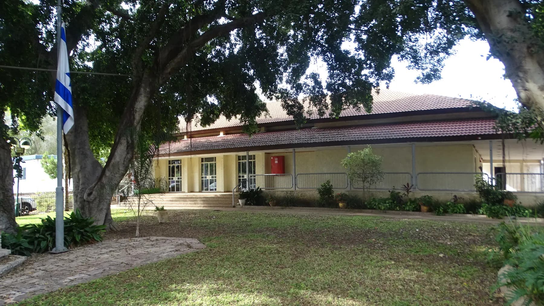 The culture house of Ramot HaShavim, a long low building with a tiled roof and a veranda, behind a lawn, a flagpole and a large shade tree.
