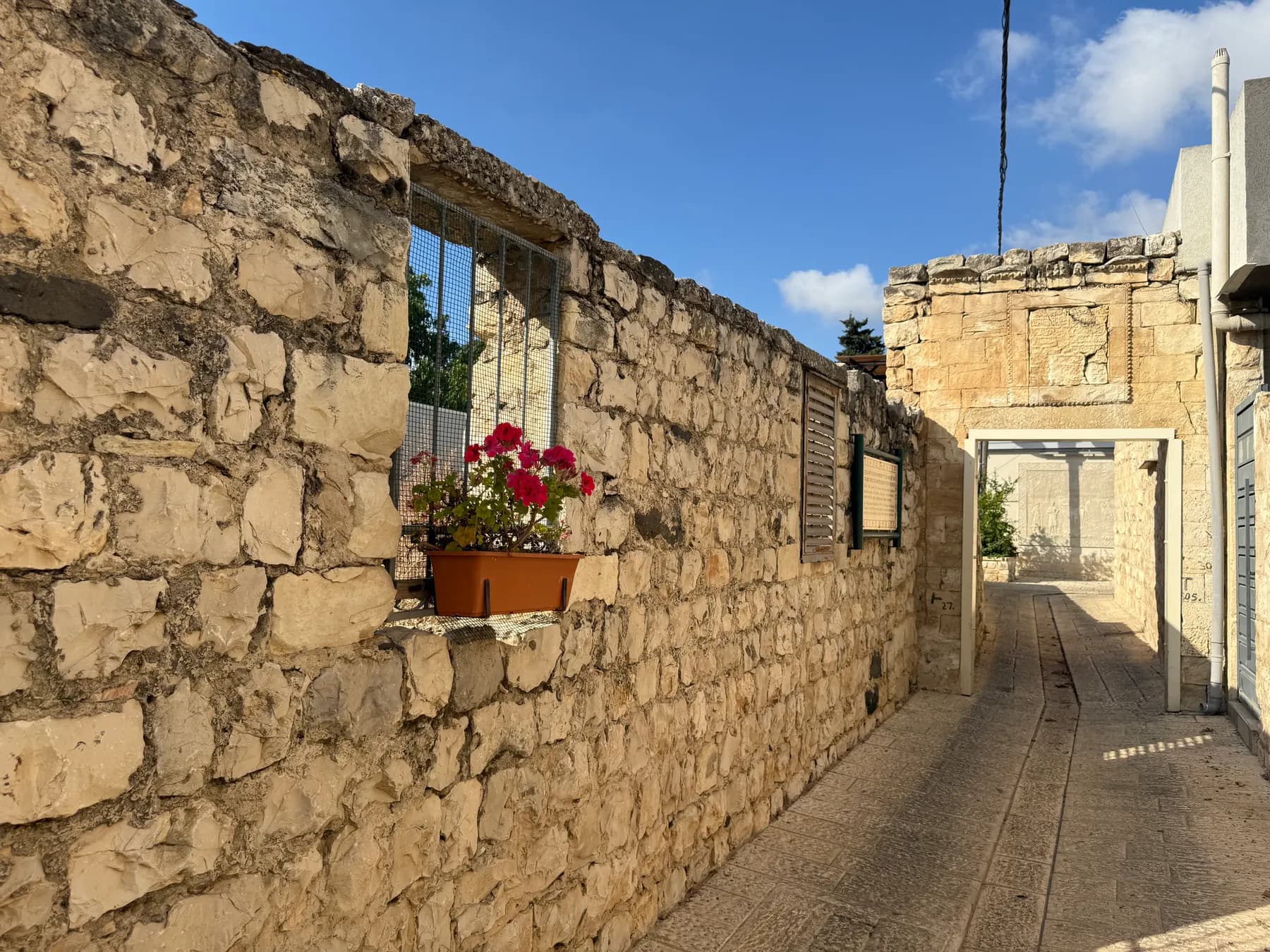 A paved lane between old stone walls in Rehaniya, with red geraniums in a window box and a stone house at the end of the lane.