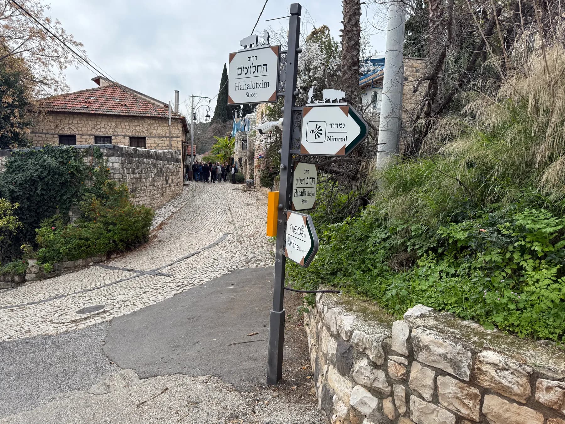 A cobblestone lane climbing between old stone houses and walls in Rosh Pinna, with street signs on a pole and people walking further up.
