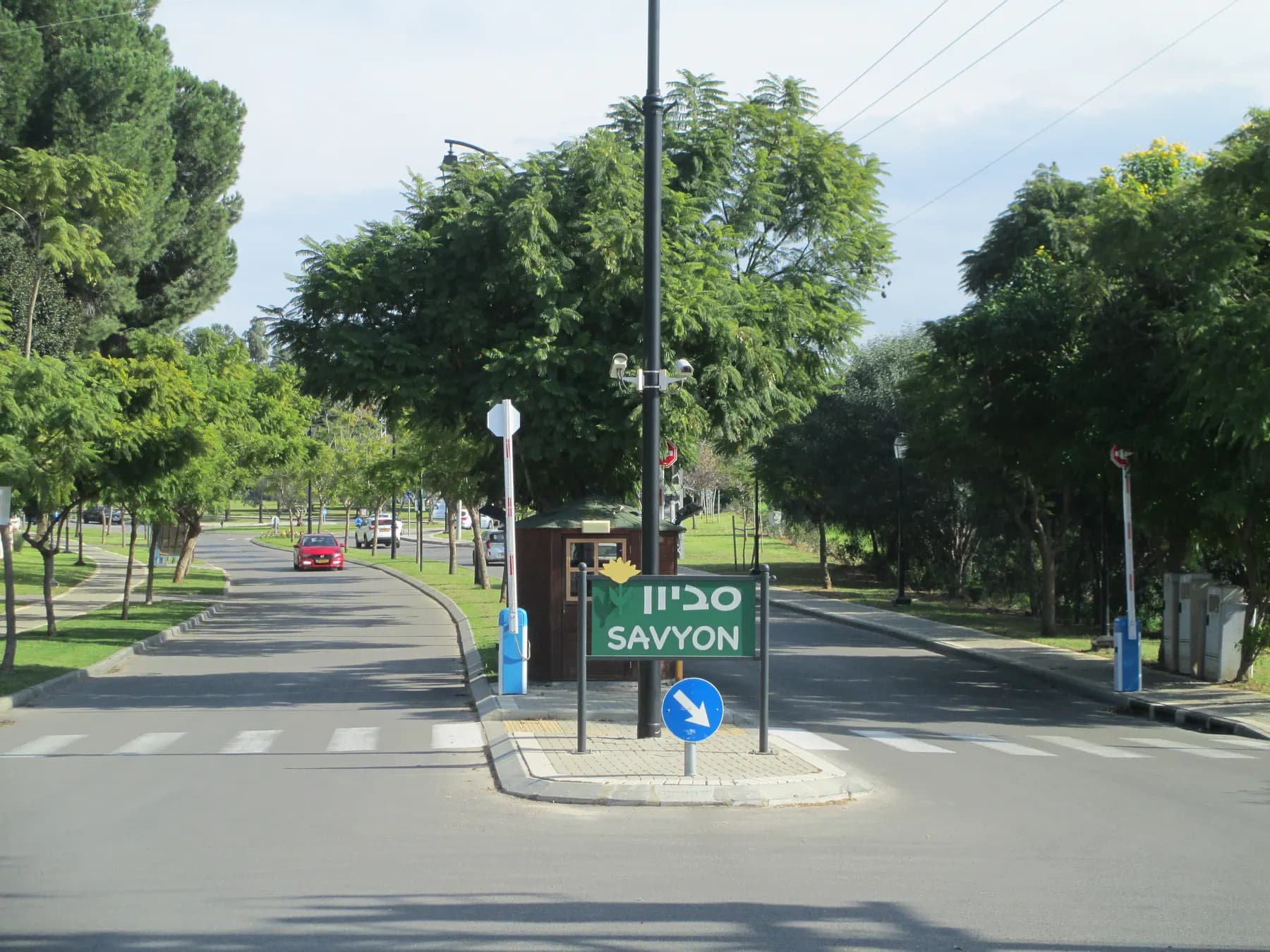 The entrance road into Savyon, lined with tall leafy trees, with a green Savyon sign on a traffic island and a gatehouse beside the road.