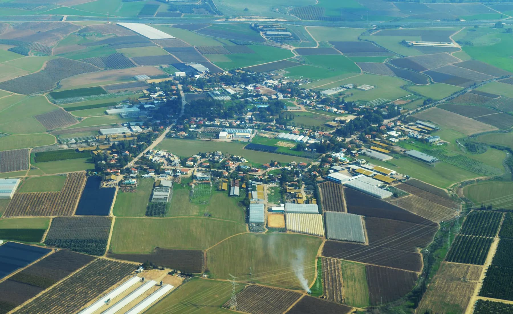 An aerial view of Sde Moshe, its houses and farm buildings along a few roads in the middle of a patchwork of green and brown fields.