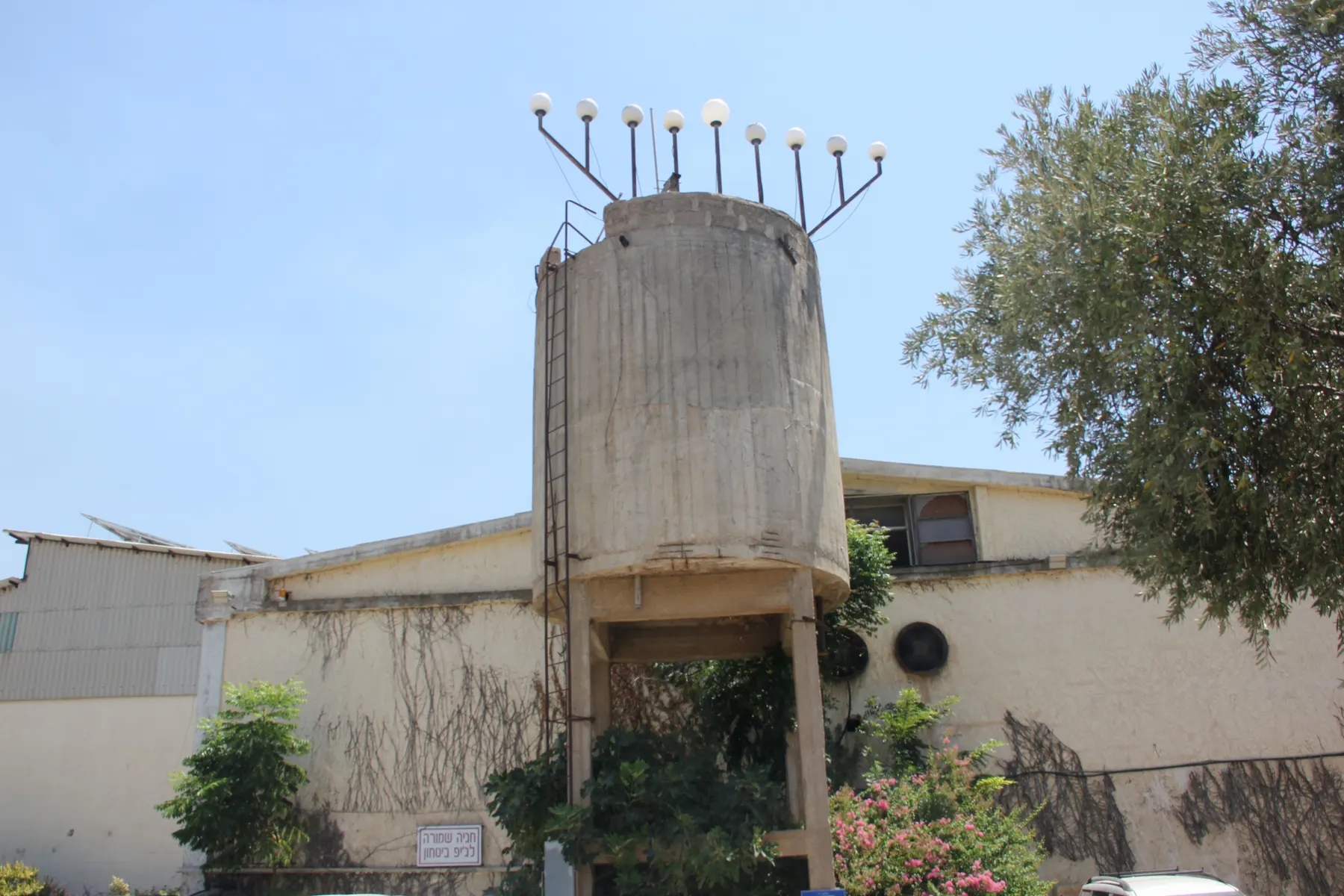 A small concrete water tower topped with a Hanukkah menorah of globe lamps in Sde Nehemia, beside a low building and an olive tree.