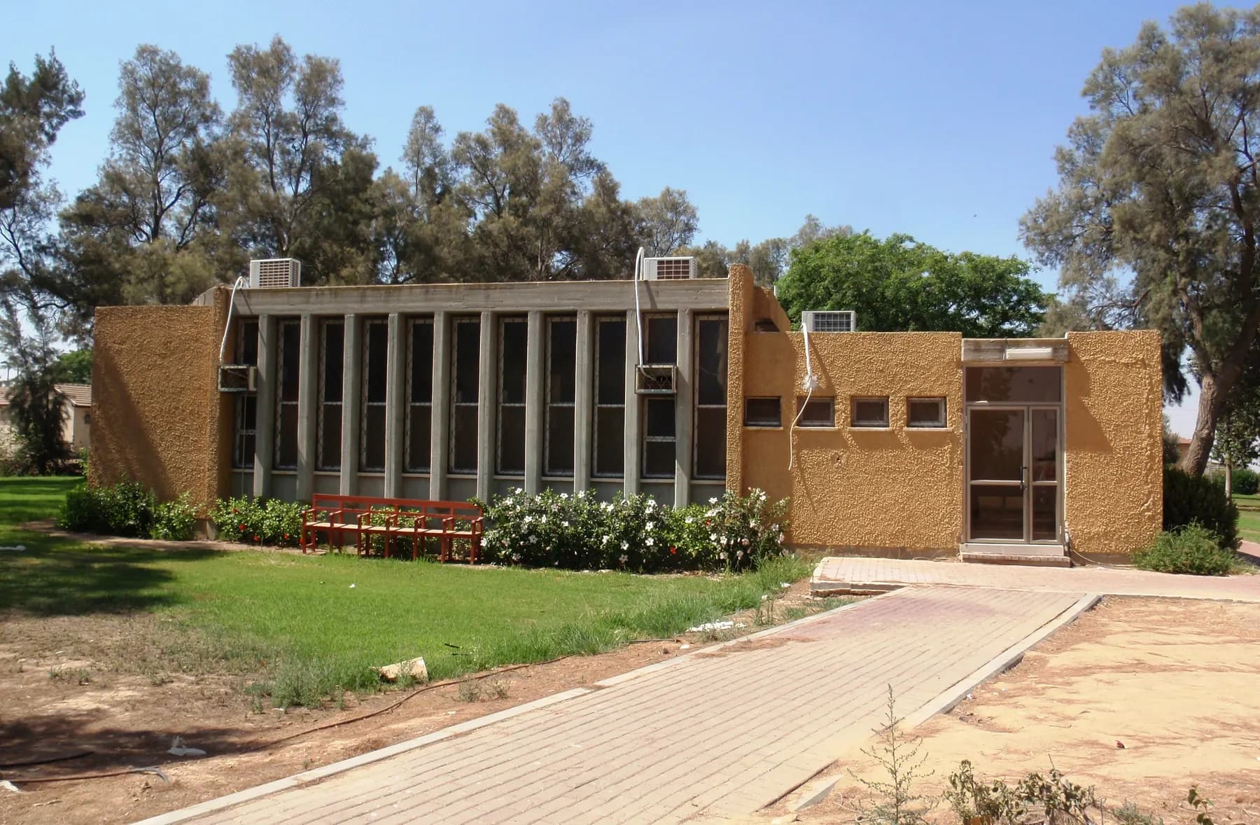 The Ohel Ya'akov synagogue in Sde Tzvi, an ochre building with a row of tall narrow windows, behind a lawn, a red bench, flowering shrubs and a brick path.
