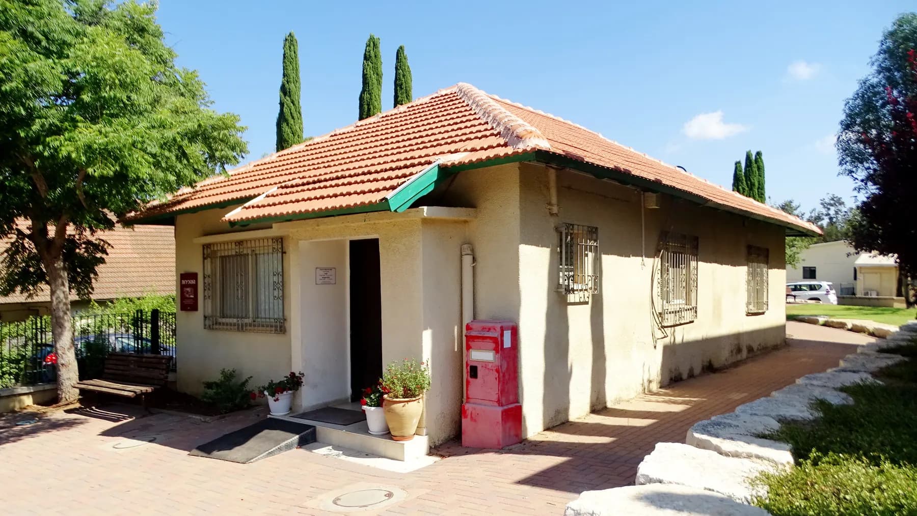 The secretariat building of Sde Warburg, a small cream-coloured house with a tiled roof, potted plants and an old red postbox by its door, with cypress trees behind.